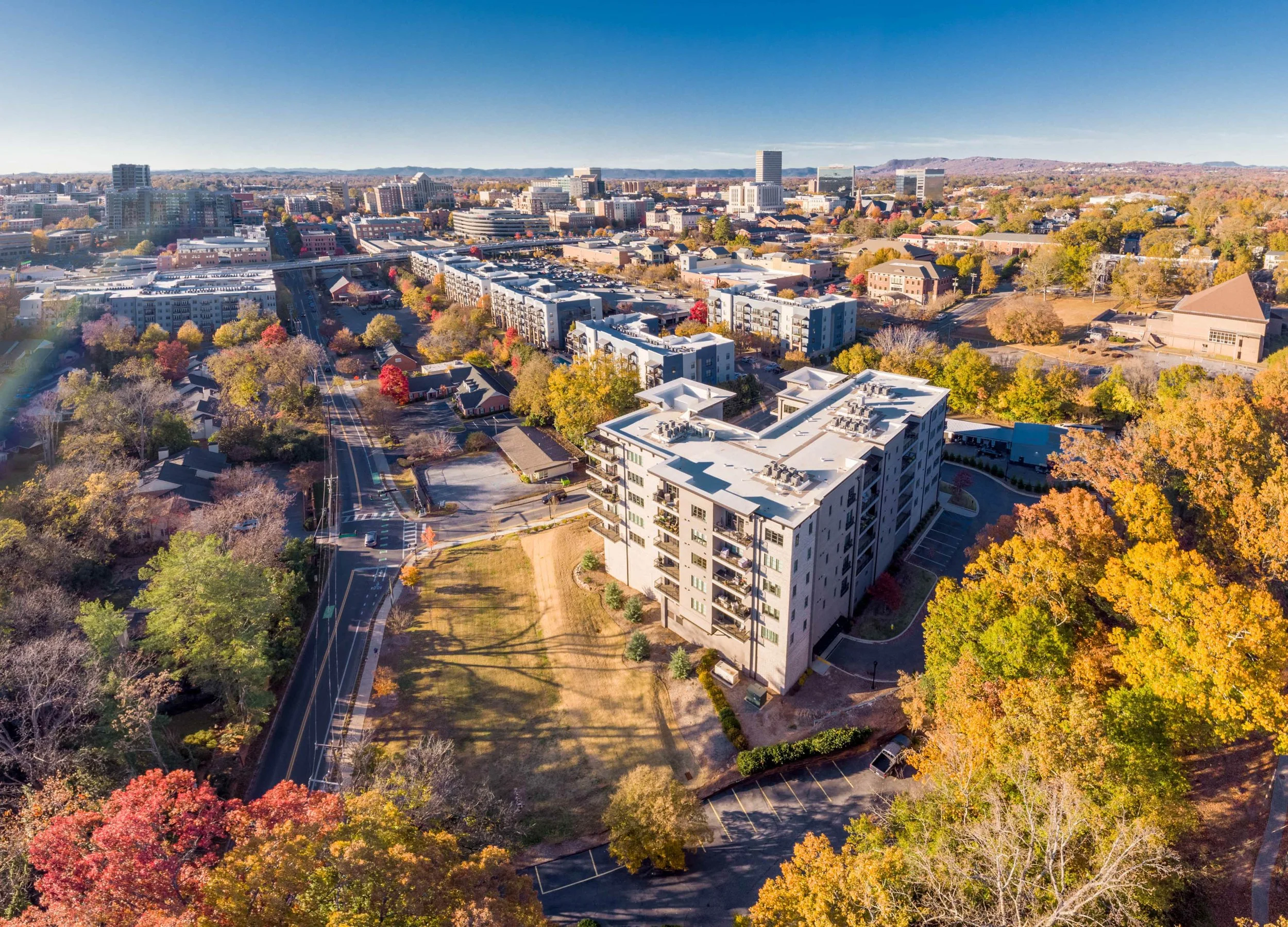 A drone photograph of luxury condominiums in Greenville, SC, with colorful fall trees, residential buildings, and a view of the Blueridge mountains and Greenville's skyline.