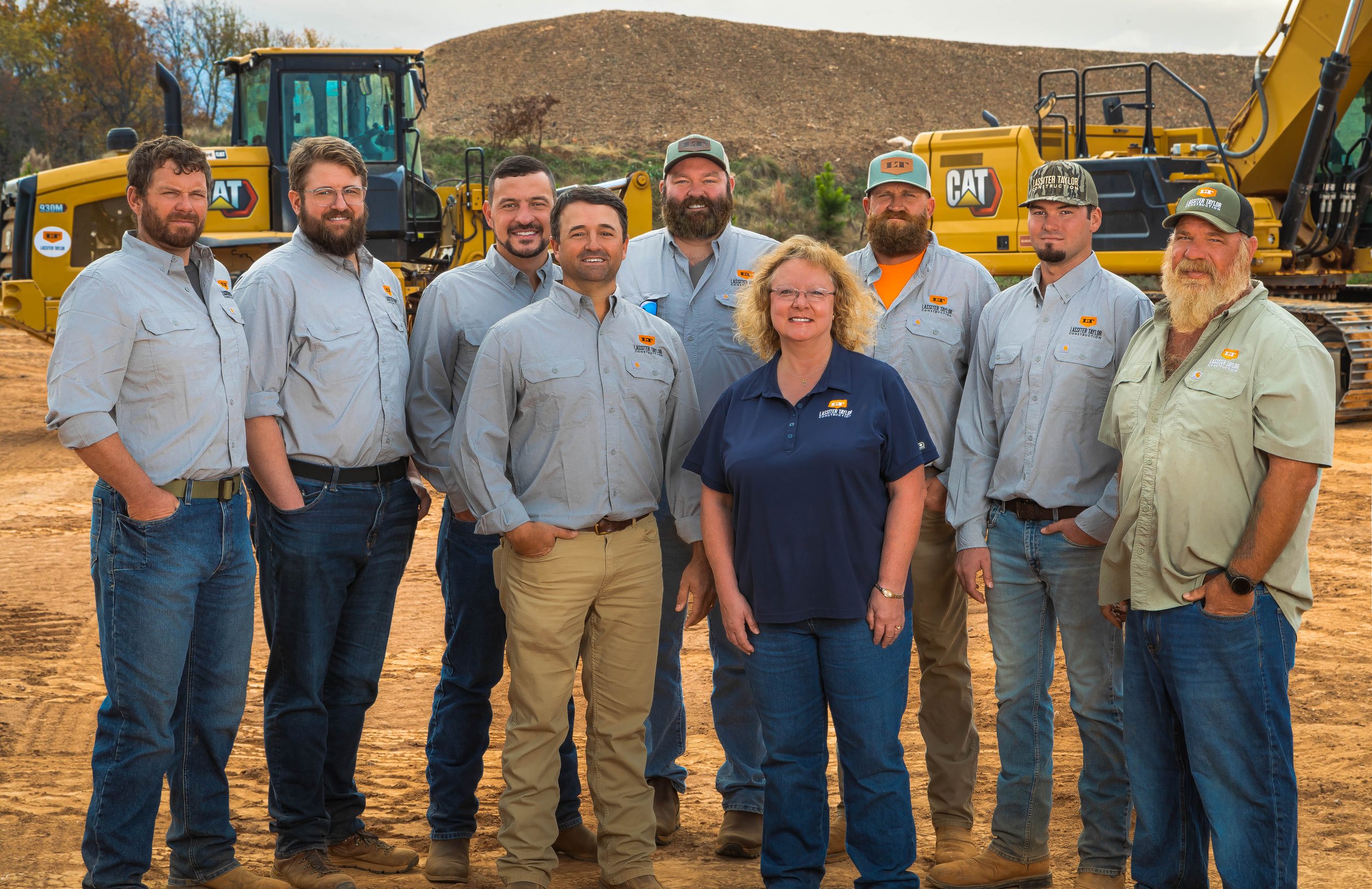 A group of nine construction workers, including eight men and one woman, standing outdoors in front of large yellow Caterpillar machinery on a dirt construction site with a hill and partly cloudy sky in the background.
