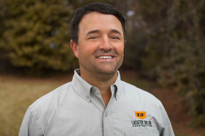 Portrait of a smiling man wearing a light gray collared shirt with a company logo, outdoors with blurred trees in the background.