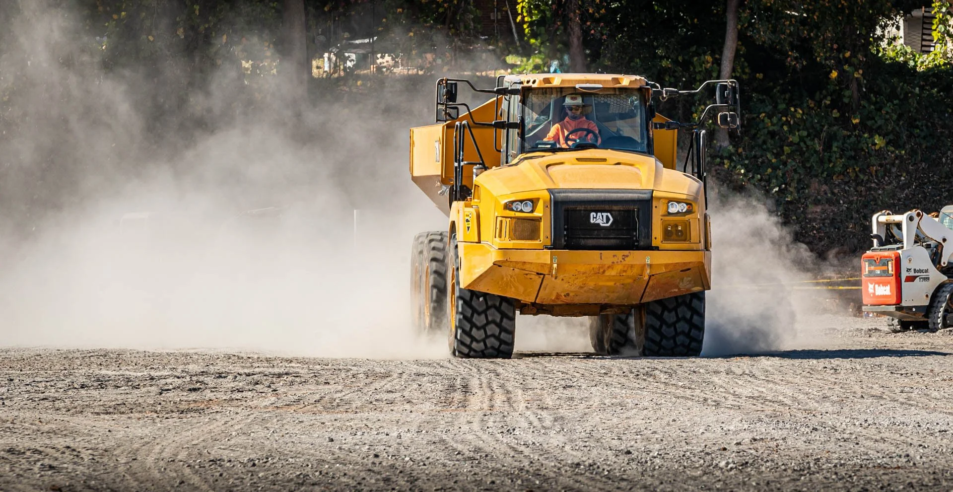 A yellow CAT construction vehicle, such as a dump truck or loader, moving on a dusty construction site with dust and dirt in the air, and construction equipment nearby.