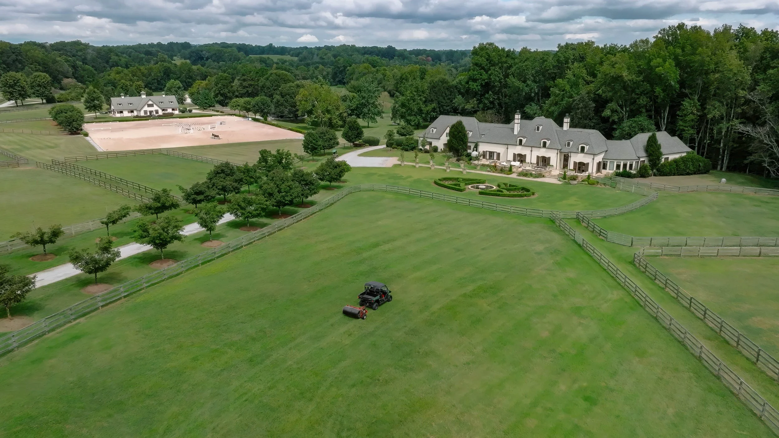 A drone photograph of a large estate with a mansion, fenced livestock areas, manicured lawn, trees, an equestrian eventing arena, and a nearby wooded area under a cloudy sky in Landrum, SC near Tryon Equestrian Center.