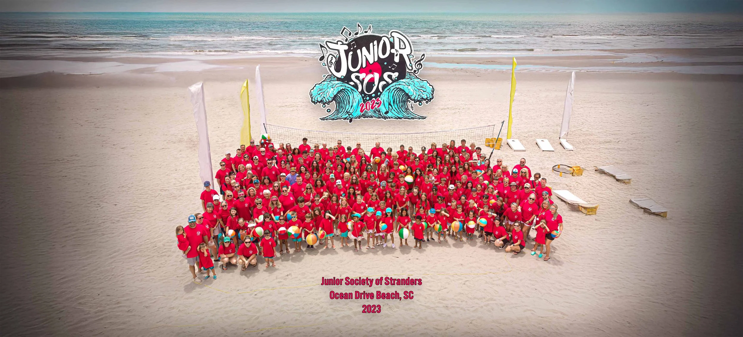 A large group of people wearing matching red shirts on the beach with volleyballs, standing in front of a volleyball net. In the background, there is a colorful banner with waves and the text "Junior SOS 2023". The text "Junior Society of Stranders O
