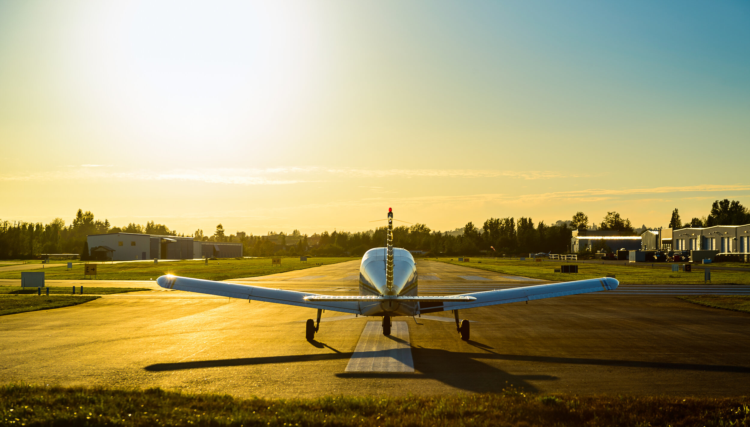 Small private airplane on runway during sunset at an airfield with hangars and trees in the background.