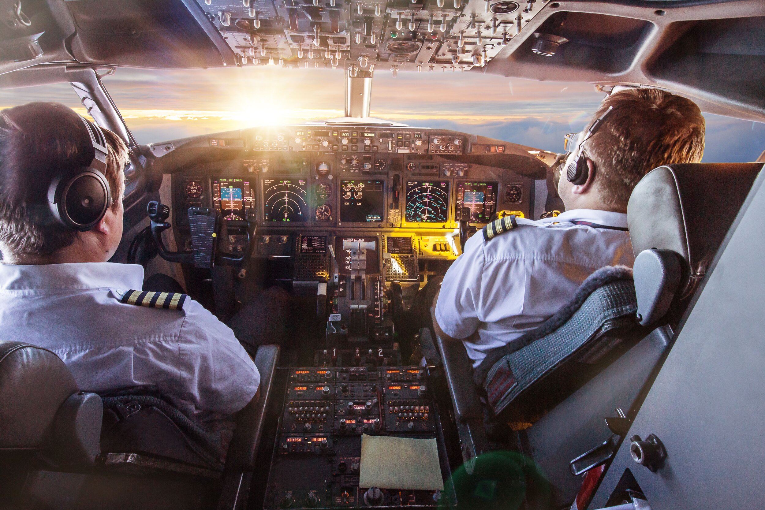 Inside the cockpit of an airplane with two pilots flying during sunset, with various instruments and controls visible.