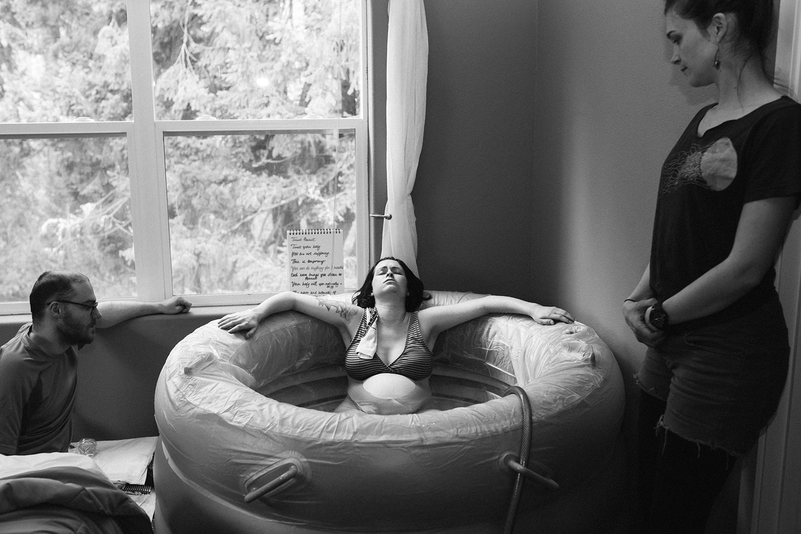 A woman in labor rests in her birthing tub at home with midwife and partner nearby.