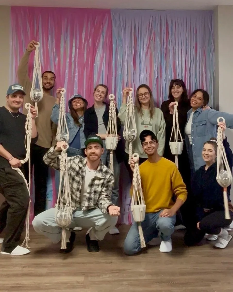Group of people at a Fun With Fibers macrame workshop, posing with their handmade macrame projects, smiling and standing in front of a pink and purple backdrop.