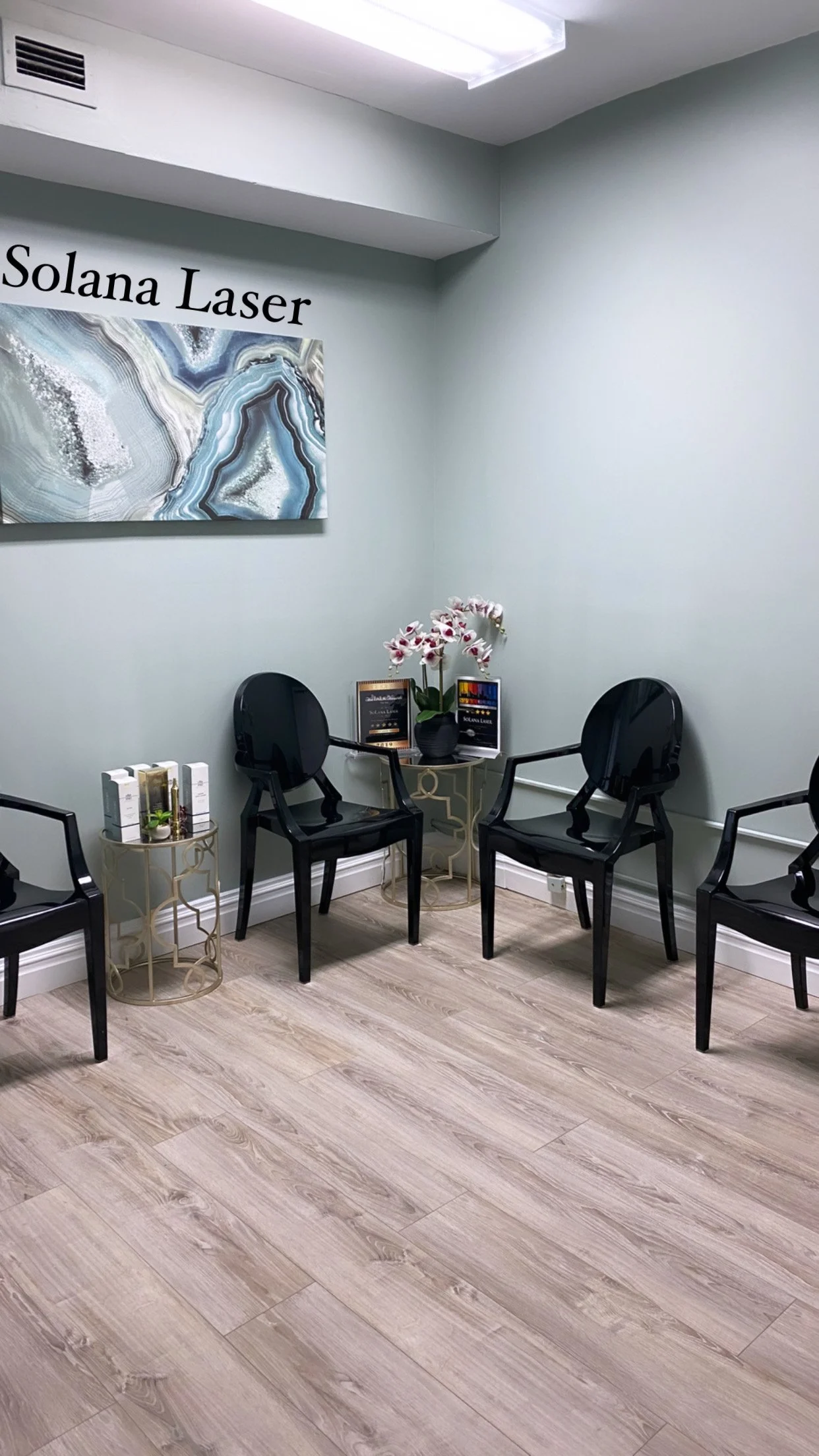Waiting room with modern black chairs, small side tables with flowers and brochures, abstract wall art, and a "Solana Laser" sign, light wood flooring, and soft green walls.