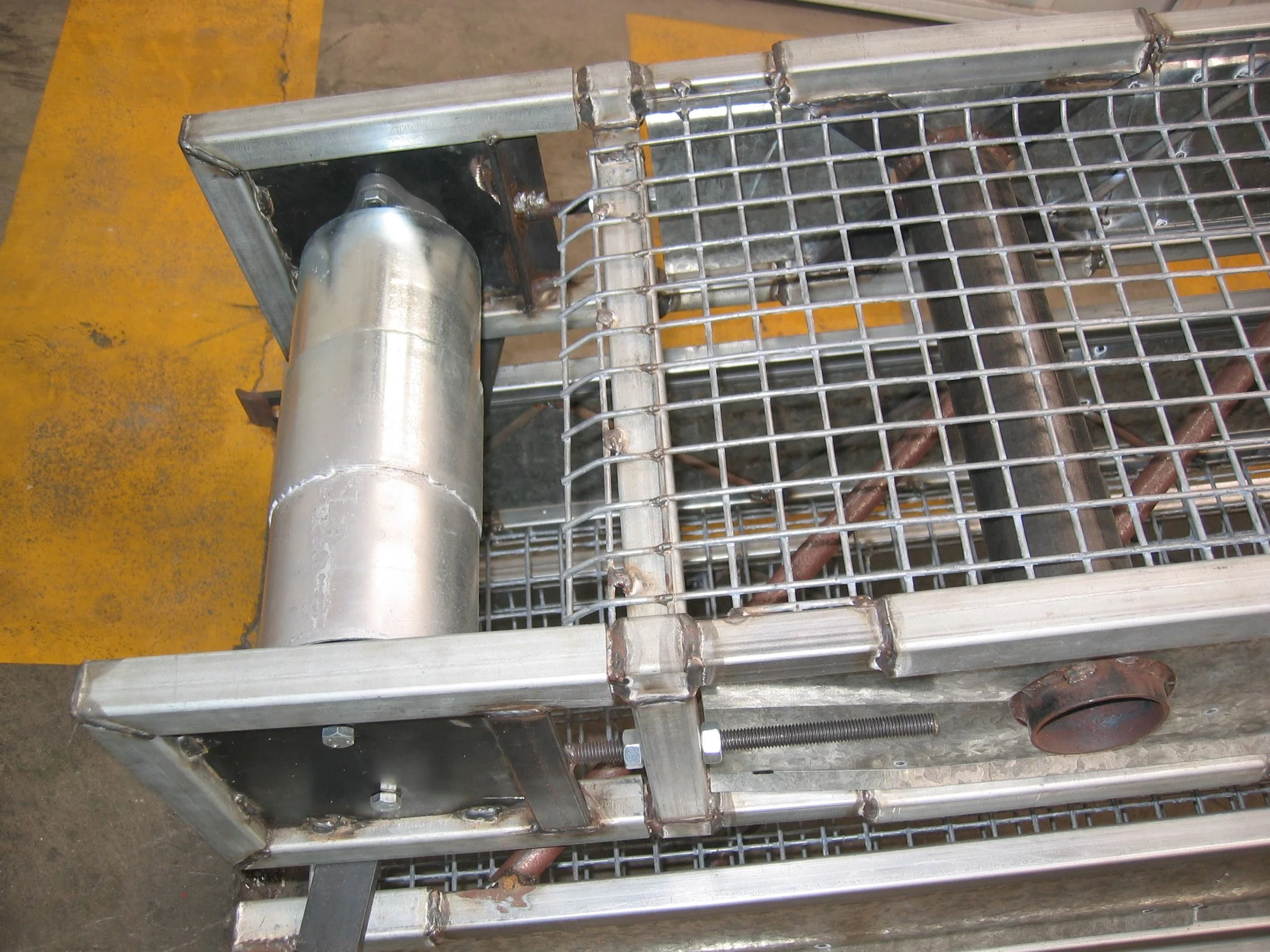 A metal cart with mesh sides and a cylindrical metal pipe on a warehouse floor with yellow safety lines.