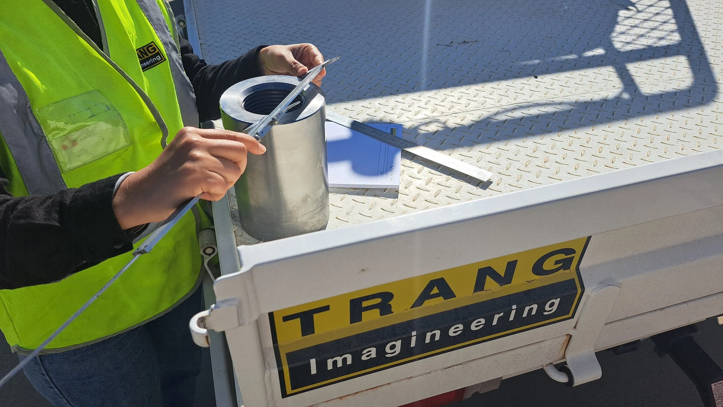 Person in a yellow safety vest inspecting a small metal pipe with a ruler and a measuring tape on a white work truck with a yellow and black 'TRANG IMAGINEERING' logo.
