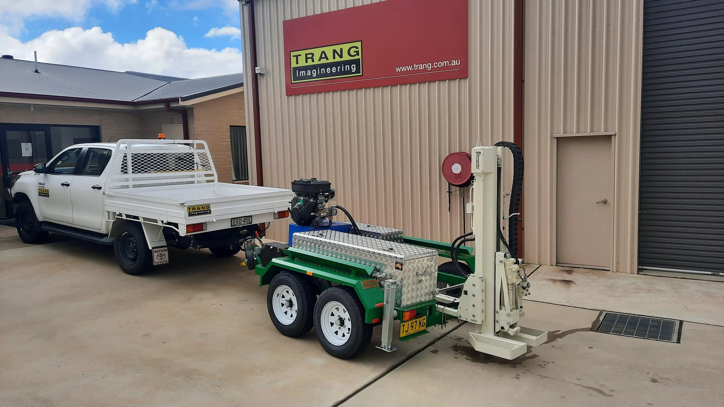 A white utility truck attached to a green and white trailer soil sampling machine with hydraulic components, parked outside a beige industrial building with a red sign that says "TRANG Imagineering".