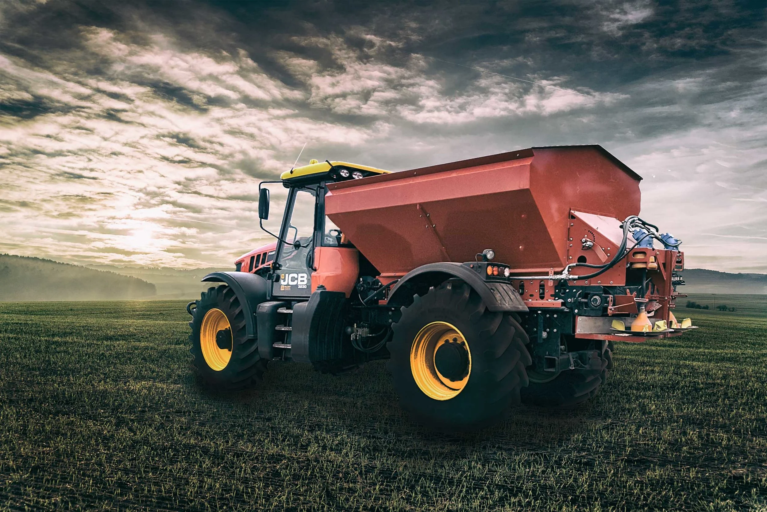Red agricultural tractor with super lime spreader in a green open field with a cloudy sky and distant hills.