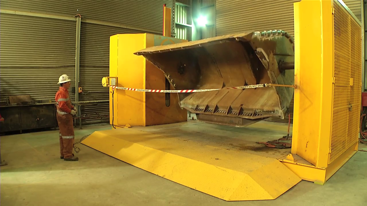 Large industrial machine with a yellow safety cover and a suspended metal bucket, inside a factory or warehouse. A worker wearing safety gear stands next to the machine.