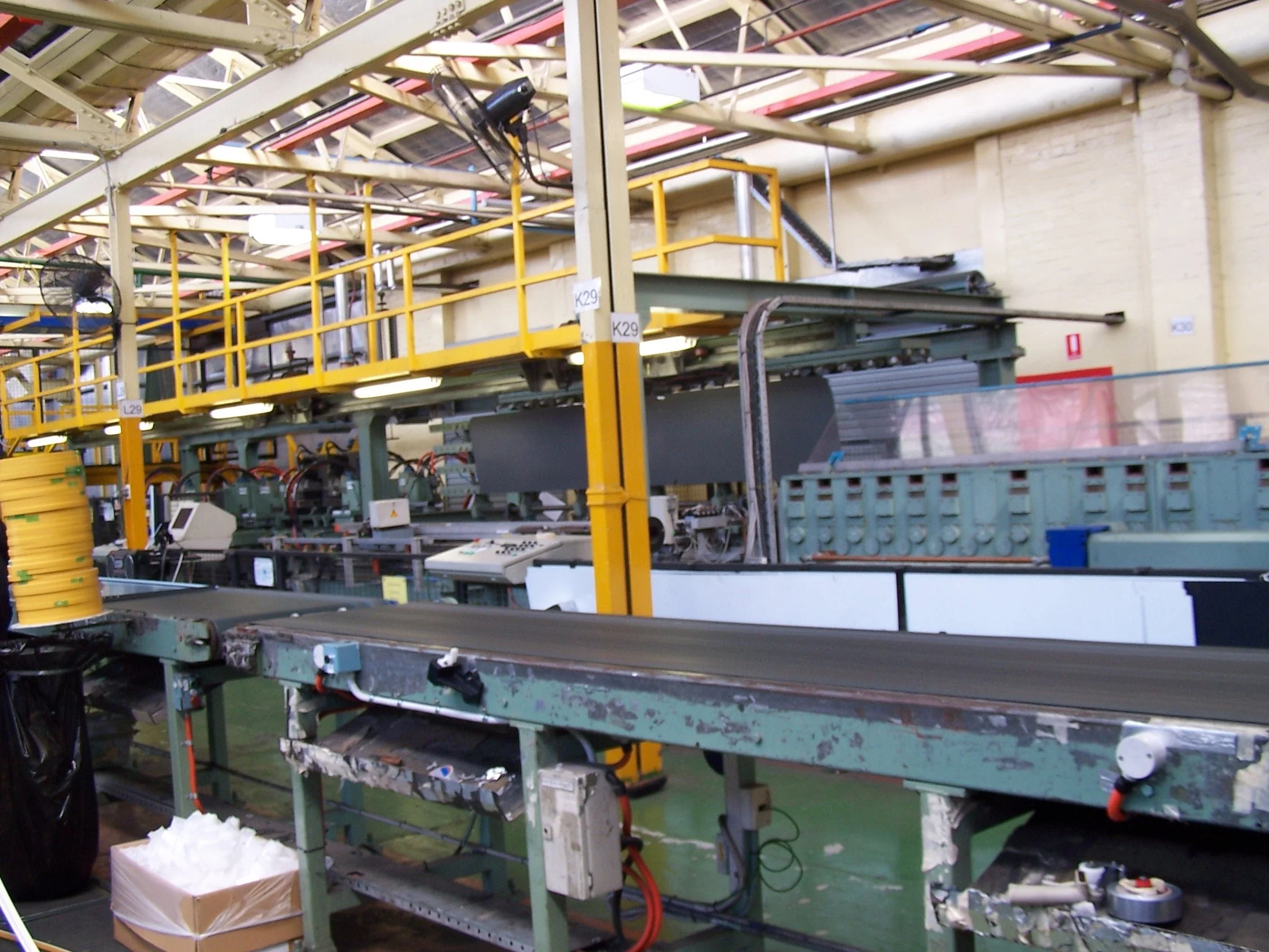 Industrial factory setting with green metal conveyor belt and various machinery, wires, and control panels. Yellow safety railing and some scattered paper and packing materials on the floor.