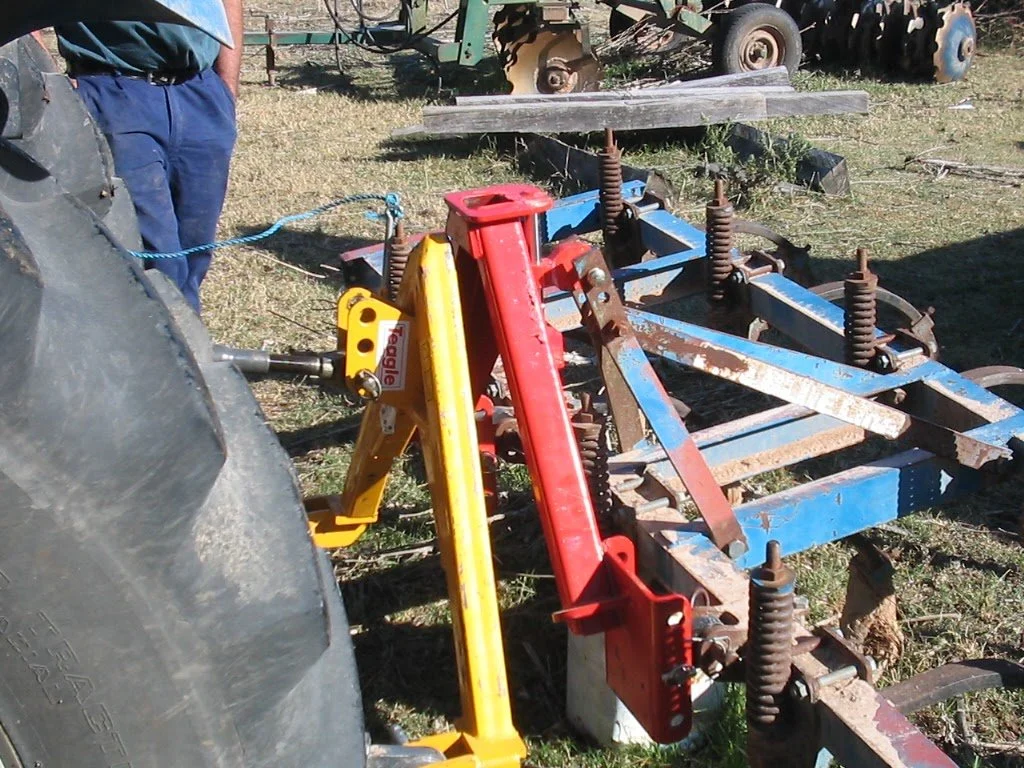 Farm equipment and machinery on grass, including a red and yellow attachment connected to a tractor, with rusty blue-colored frames and large coil springs.