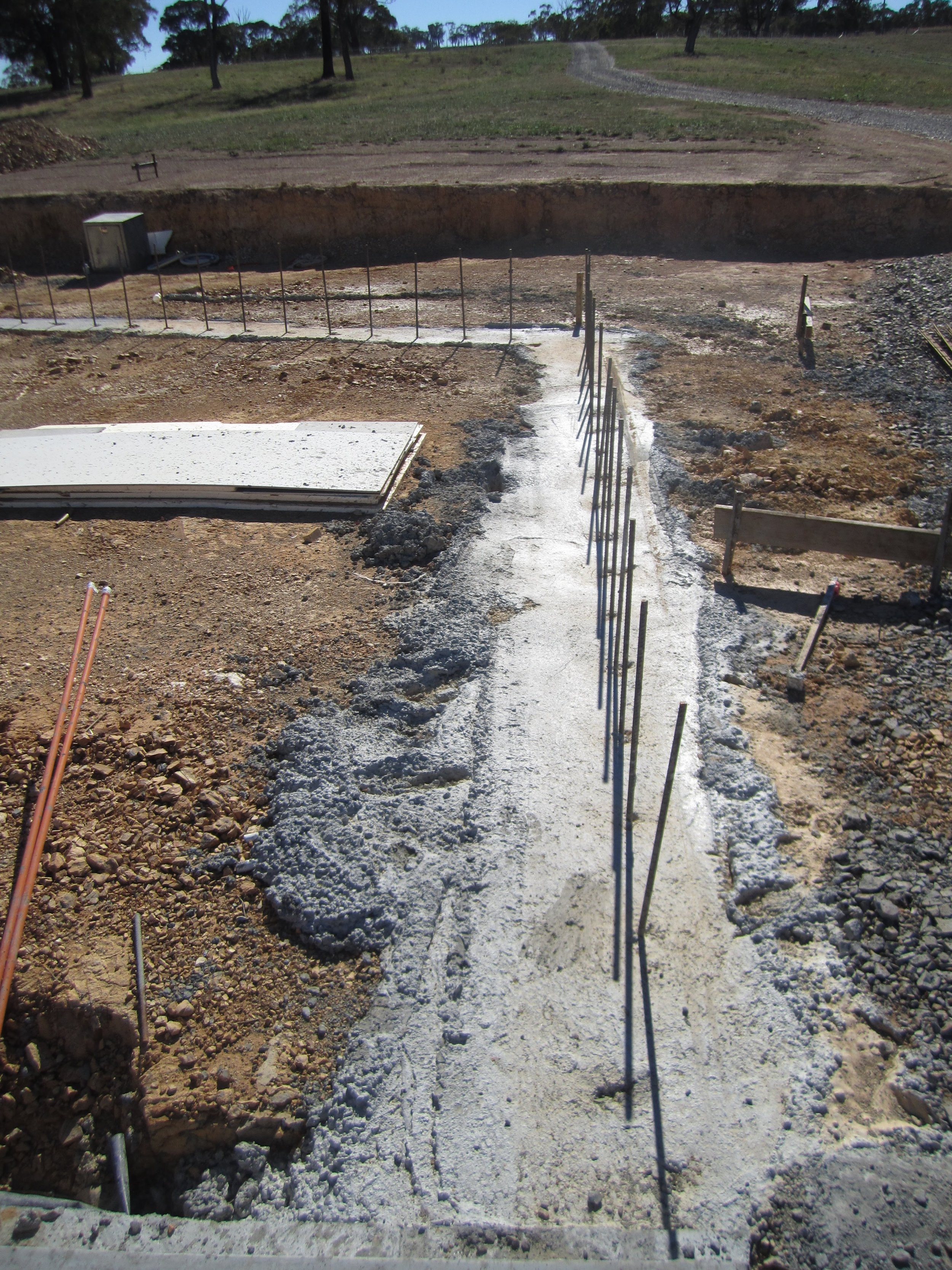 Construction site with a freshly poured concrete sidewalk, metal rebar sticks, and wooden forms, surrounded by dirt and gravel.
