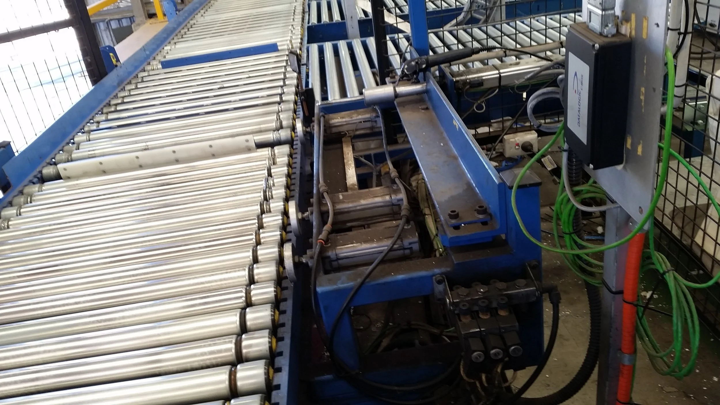 Industrial conveyor system with multiple metal rollers and blue framework in a manufacturing environment.