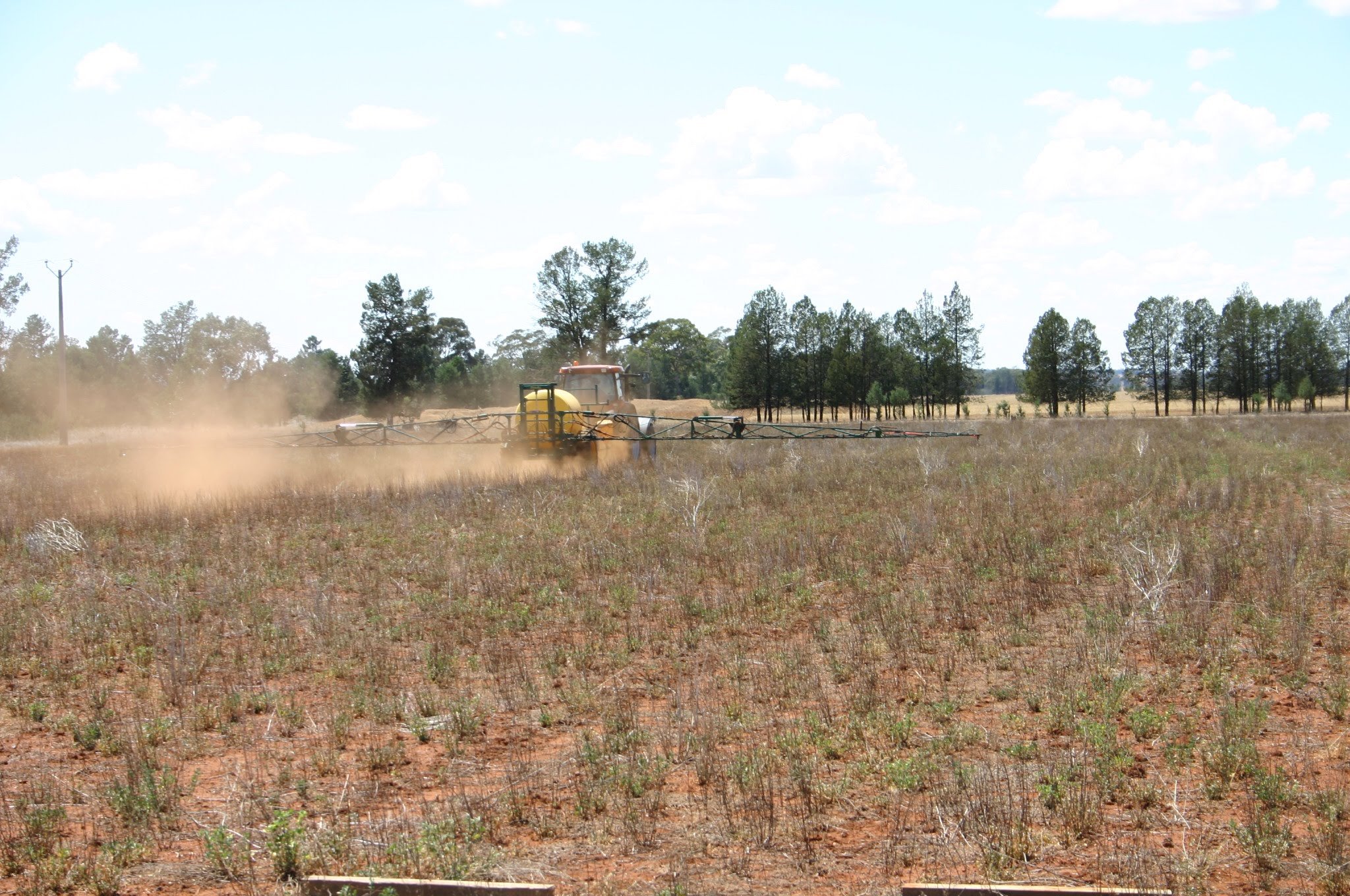 A tractor spraying pesticides or herbicides over a dry, weeded field with sparse vegetation, in a rural area with trees and power poles in the background.