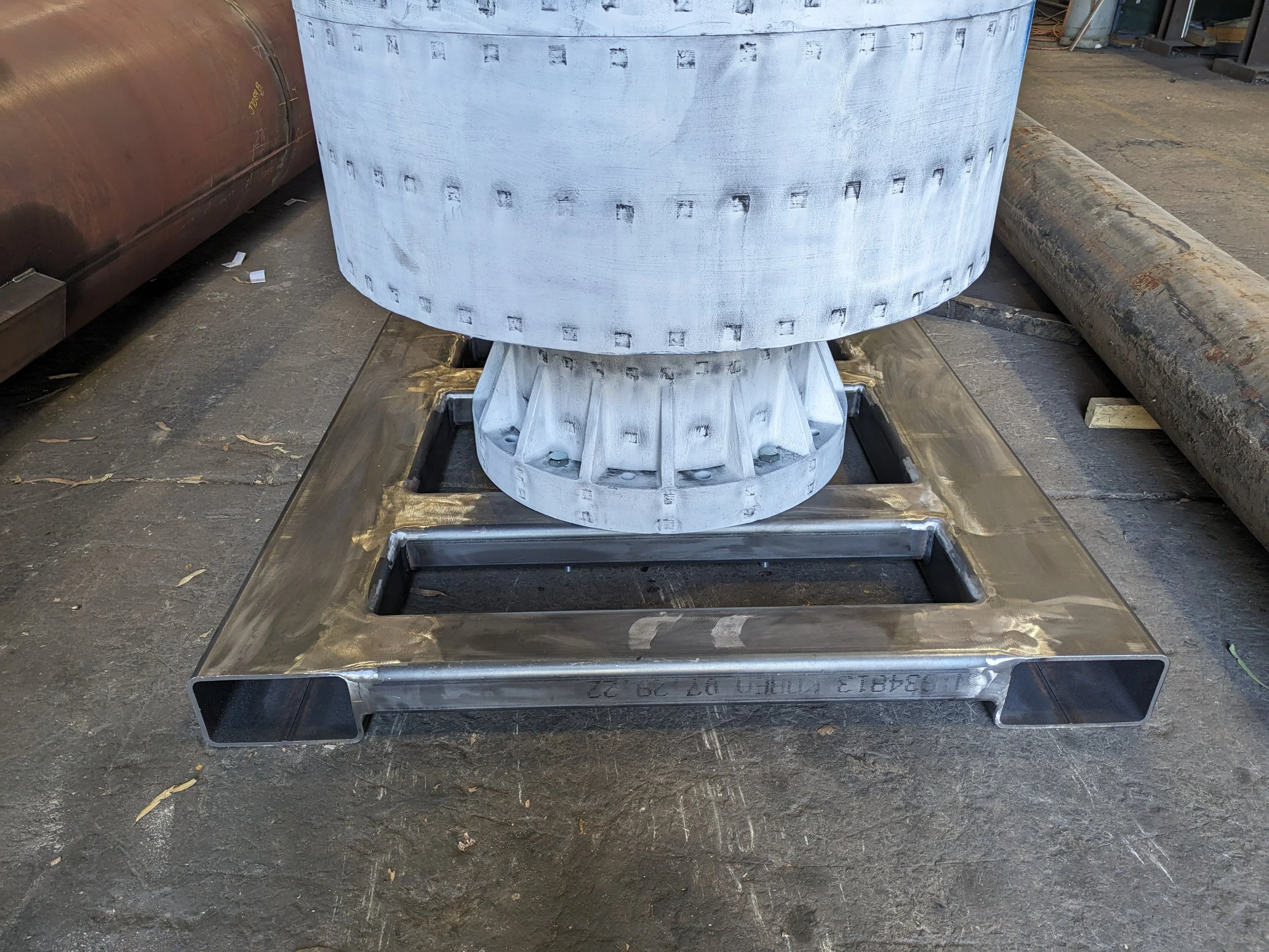 Large metallic industrial machine part with a hollow cylindrical shape and square-shaped attached base, placed on a metal platform in a workshop or factory setting.