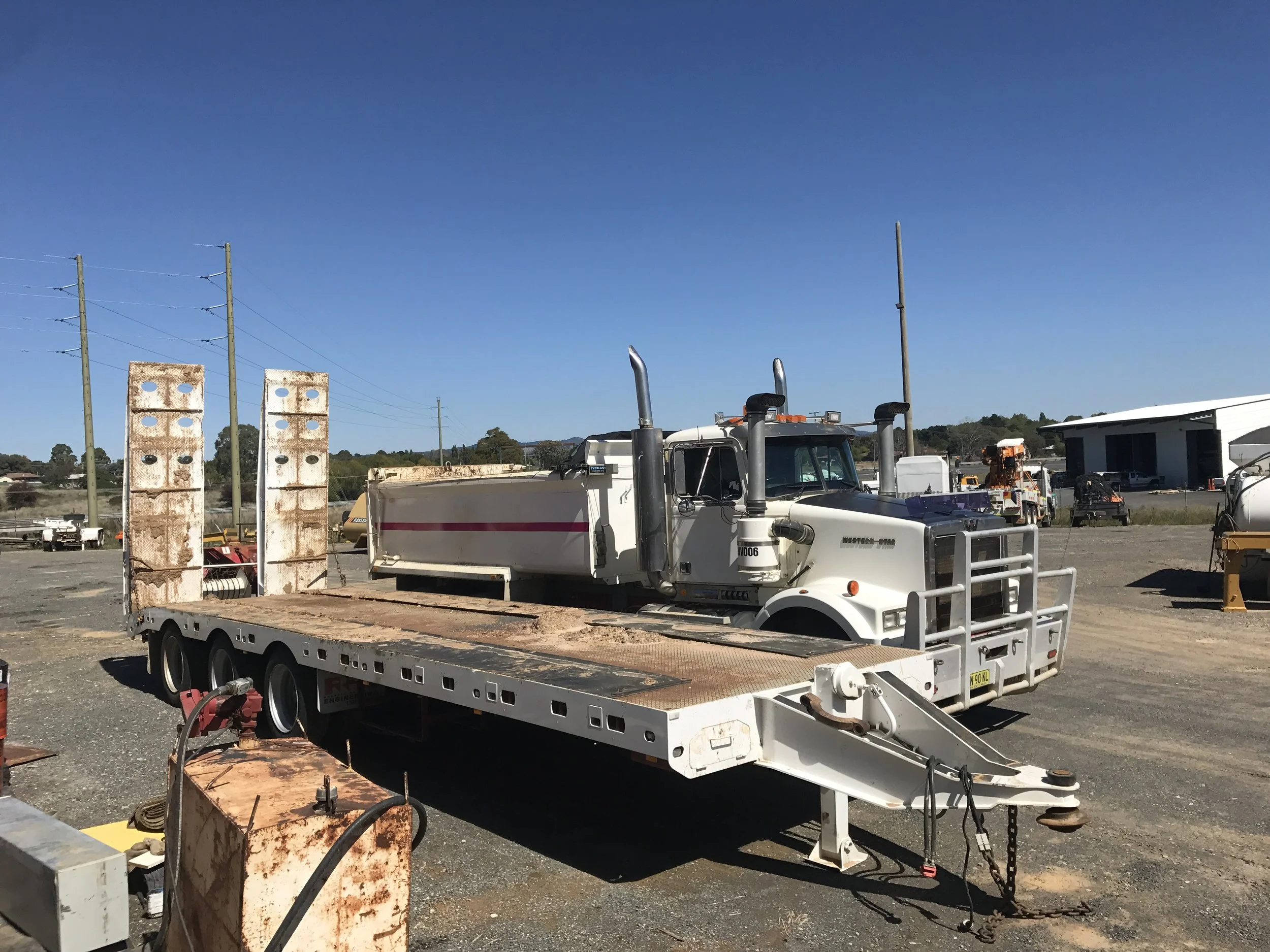 Flatbed trailer with a white cab parked on a gravel lot, surrounded by industrial equipment and other vehicles, under a clear blue sky.
