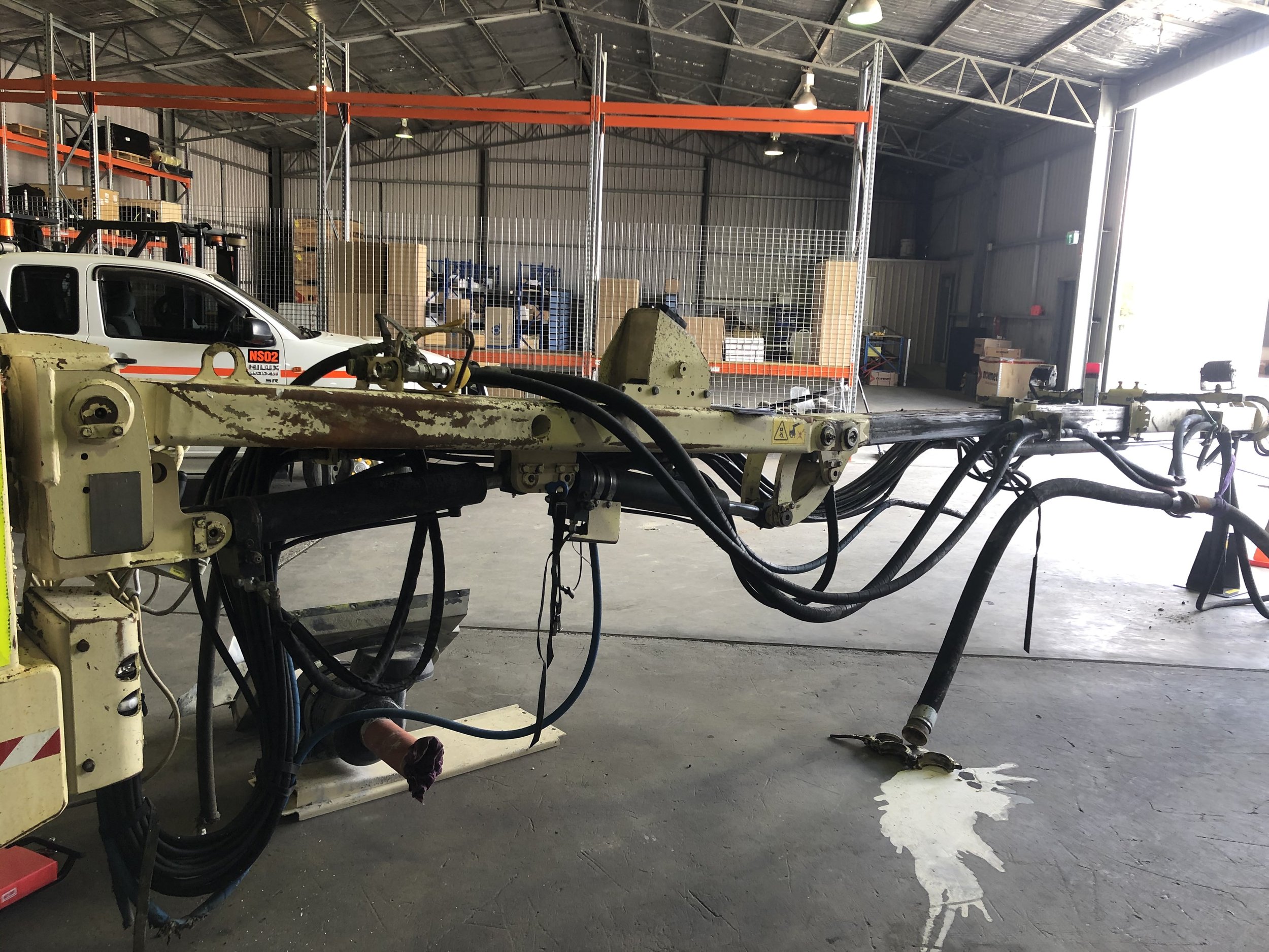 Empty industrial warehouse with a large, disassembled piece of machinery leaking oil on the concrete floor. A white vehicle with red stripe and some equipment stored on orange metal shelves in the background.
