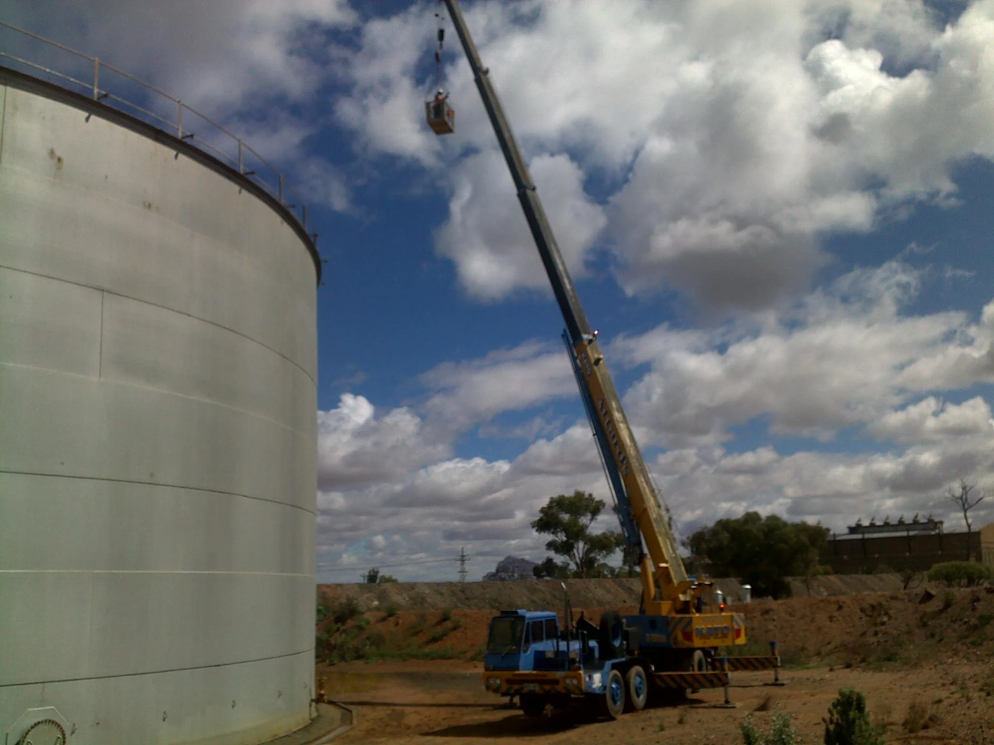 A crane truck with an extended arm lifting a worker in a basket near a large, white, cylindrical storage tank under a partly cloudy sky.