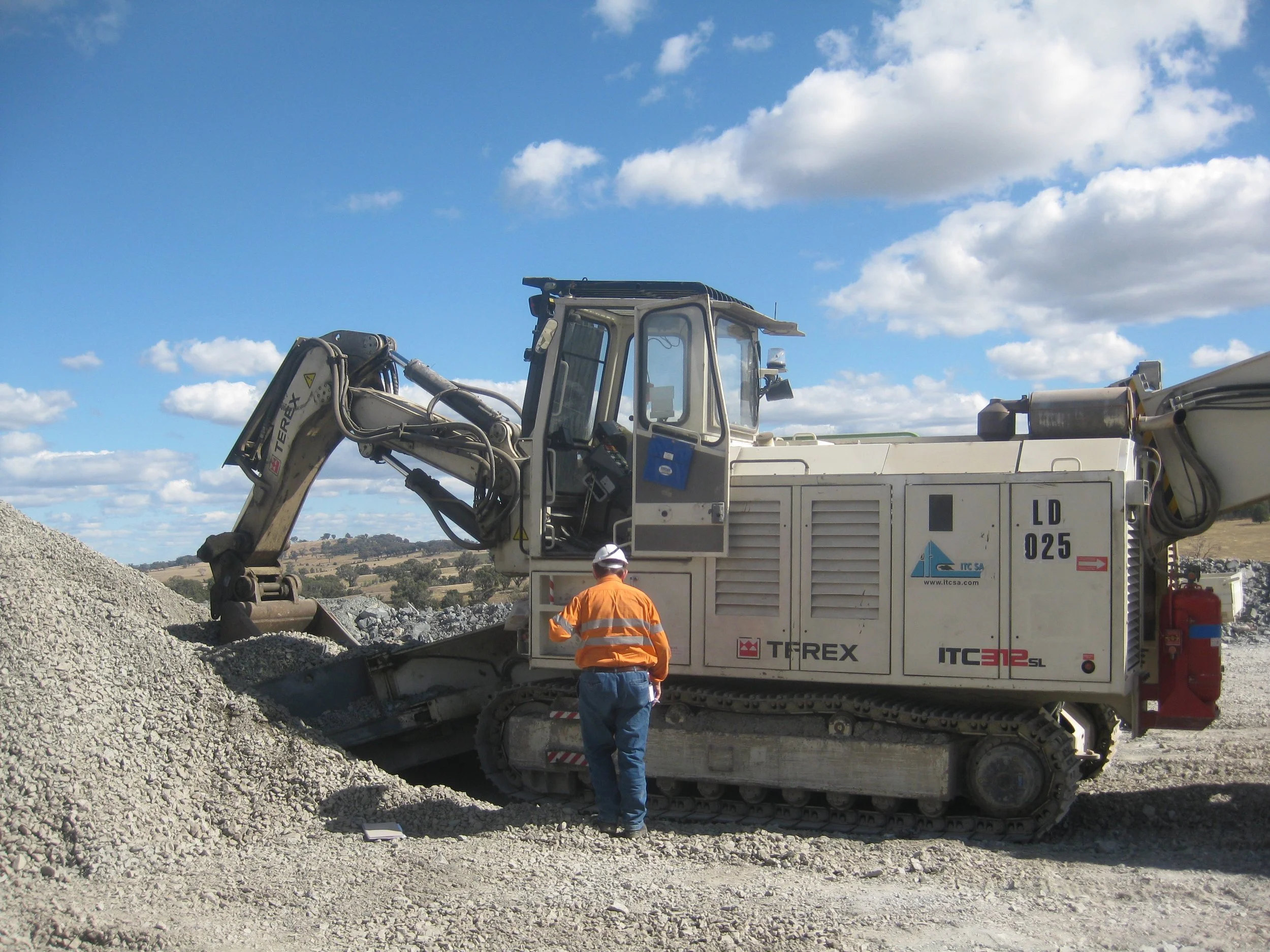 Construction worker in orange safety vest and white helmet standing next to an excavator at a construction site with a cloudy blue sky.