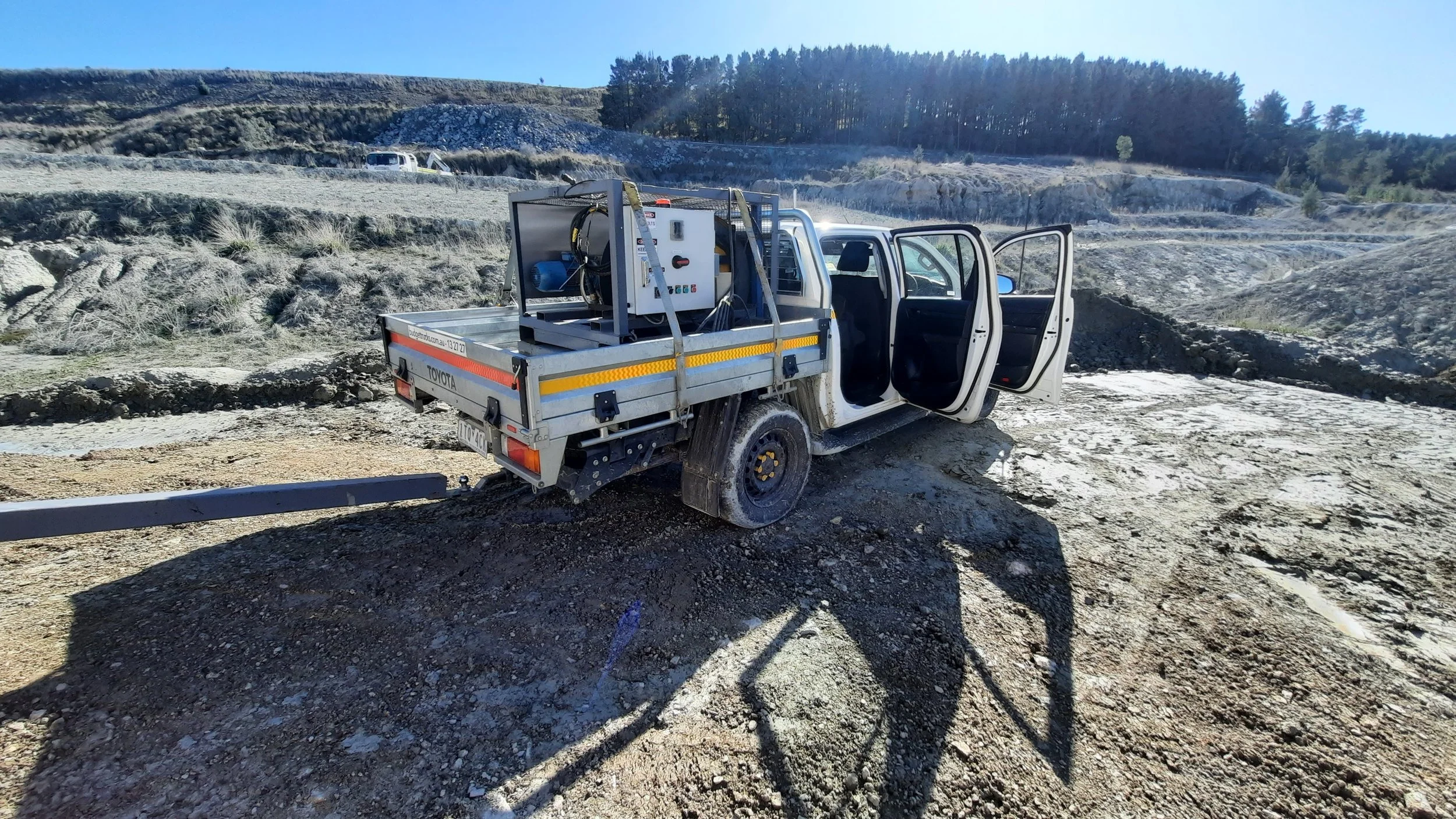 A white pickup truck with open doors parked on uneven dirt ground in a mountainous area. The truck's bed is loaded with equipment, possibly for construction or maintenance, with hills and trees in the background under a clear blue sky.