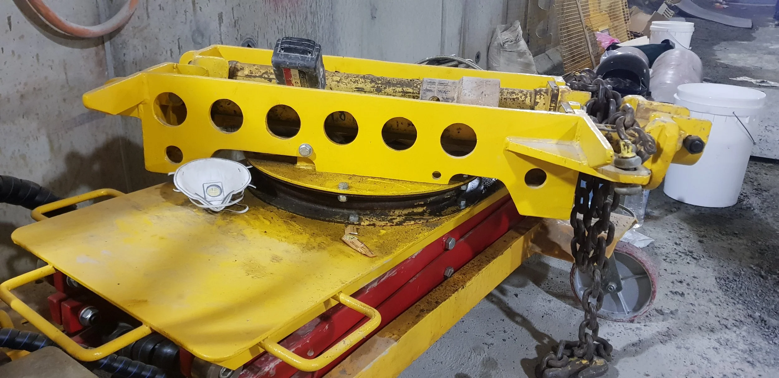 Construction equipment with a yellow frame, placed on a red base, situated in an unfinished construction site. The equipment has a large chain hanging from its side, and various tools and items are scattered around in a messy environment.