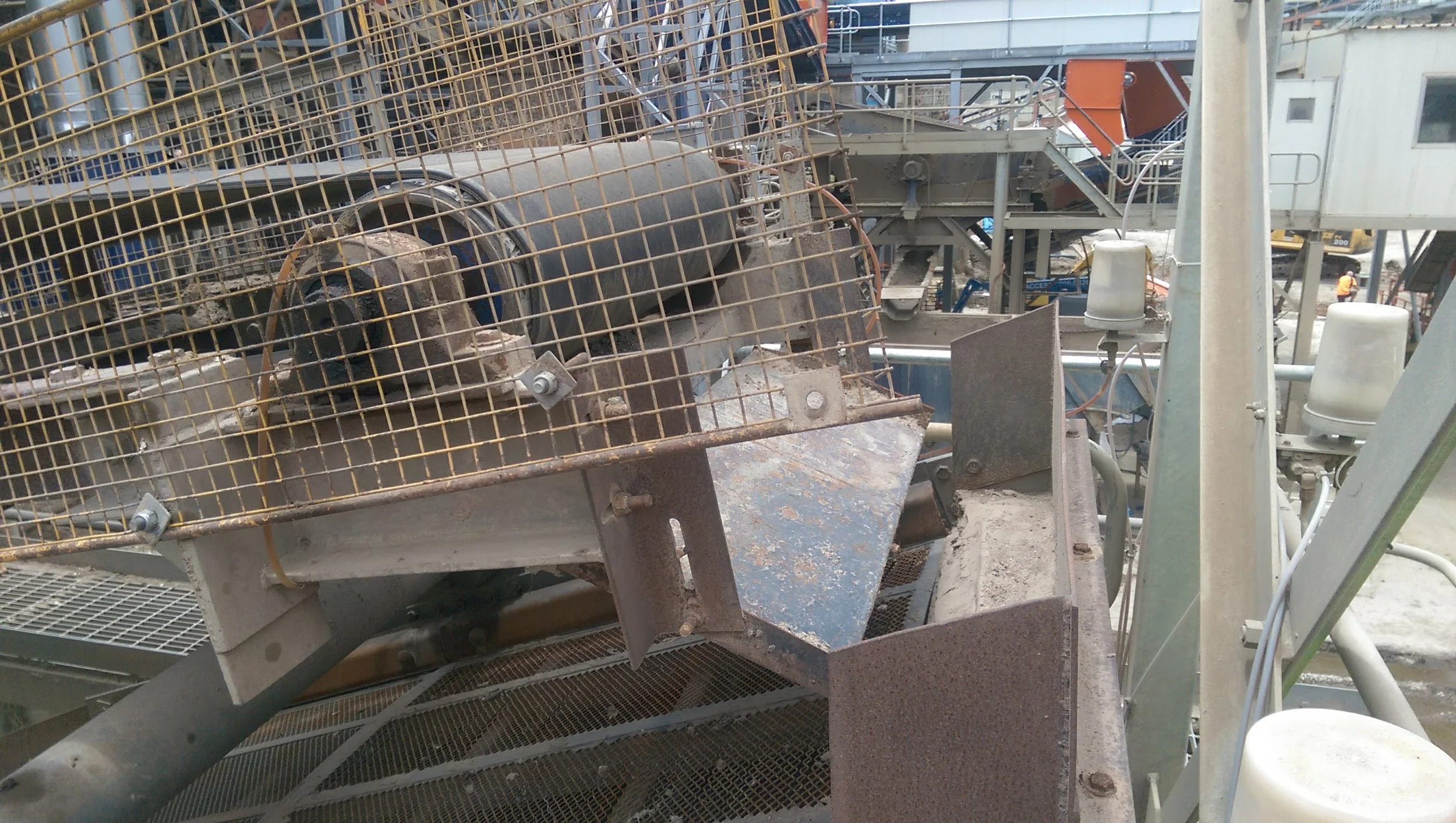 A construction site with machinery and equipment, including a large metal drum, a grated metal platform, and various pipes and structures in the background.