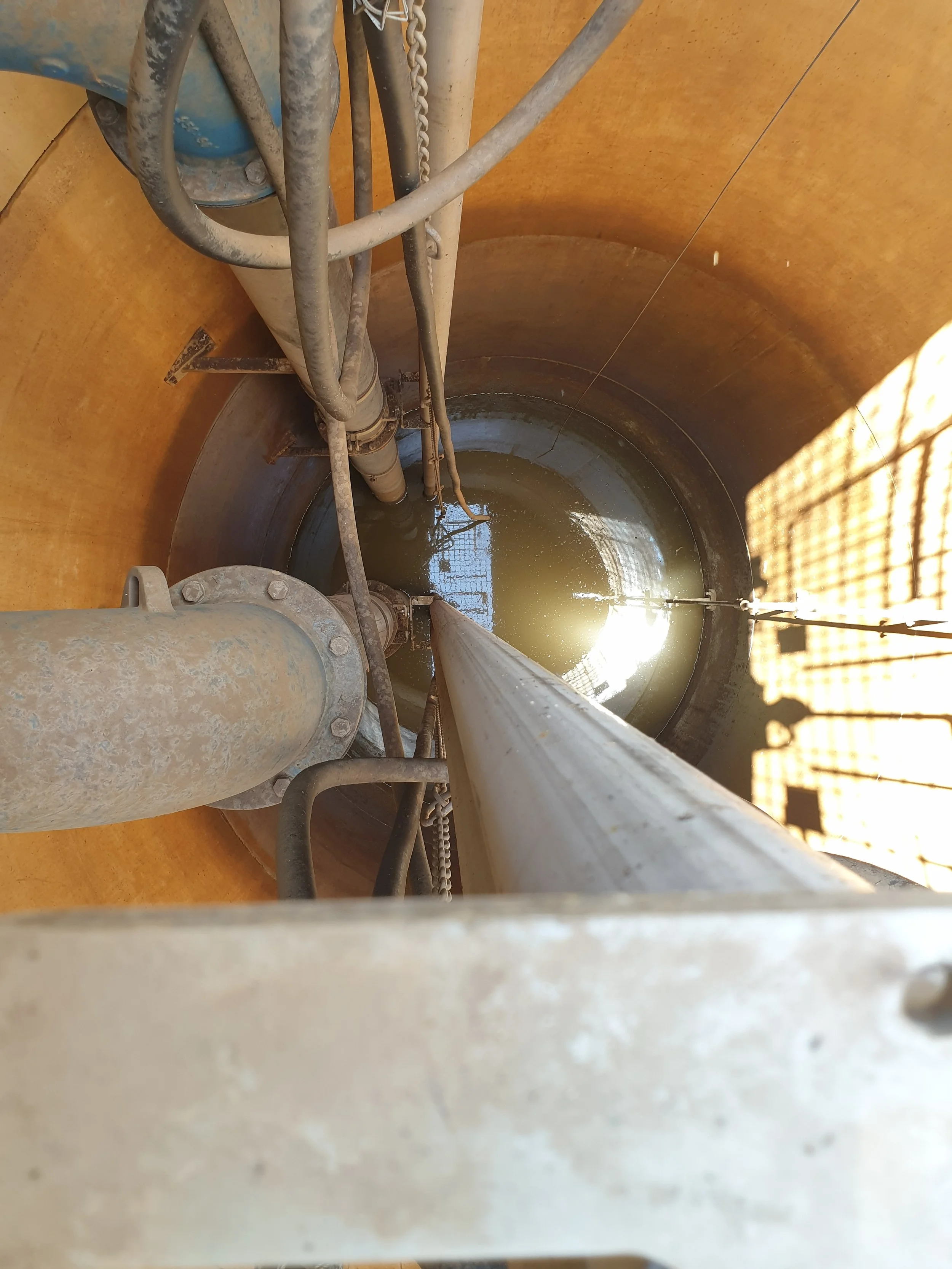 View inside a large industrial tank or well, with pipes and cables, partially filled with murky water, and a metal structure on the side.