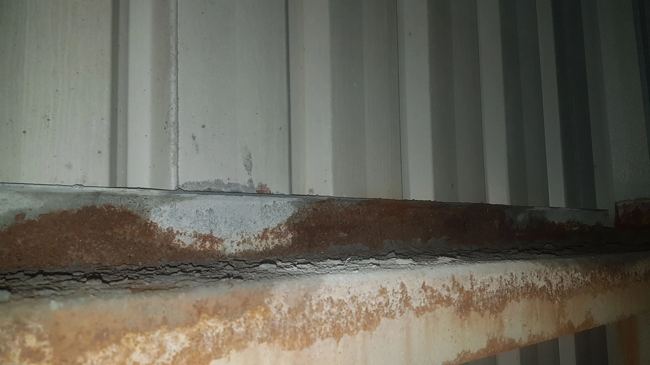 Close-up of a metal surface with visible rust and corrosion, showing a horizontal seam and vertical paneling in the background.