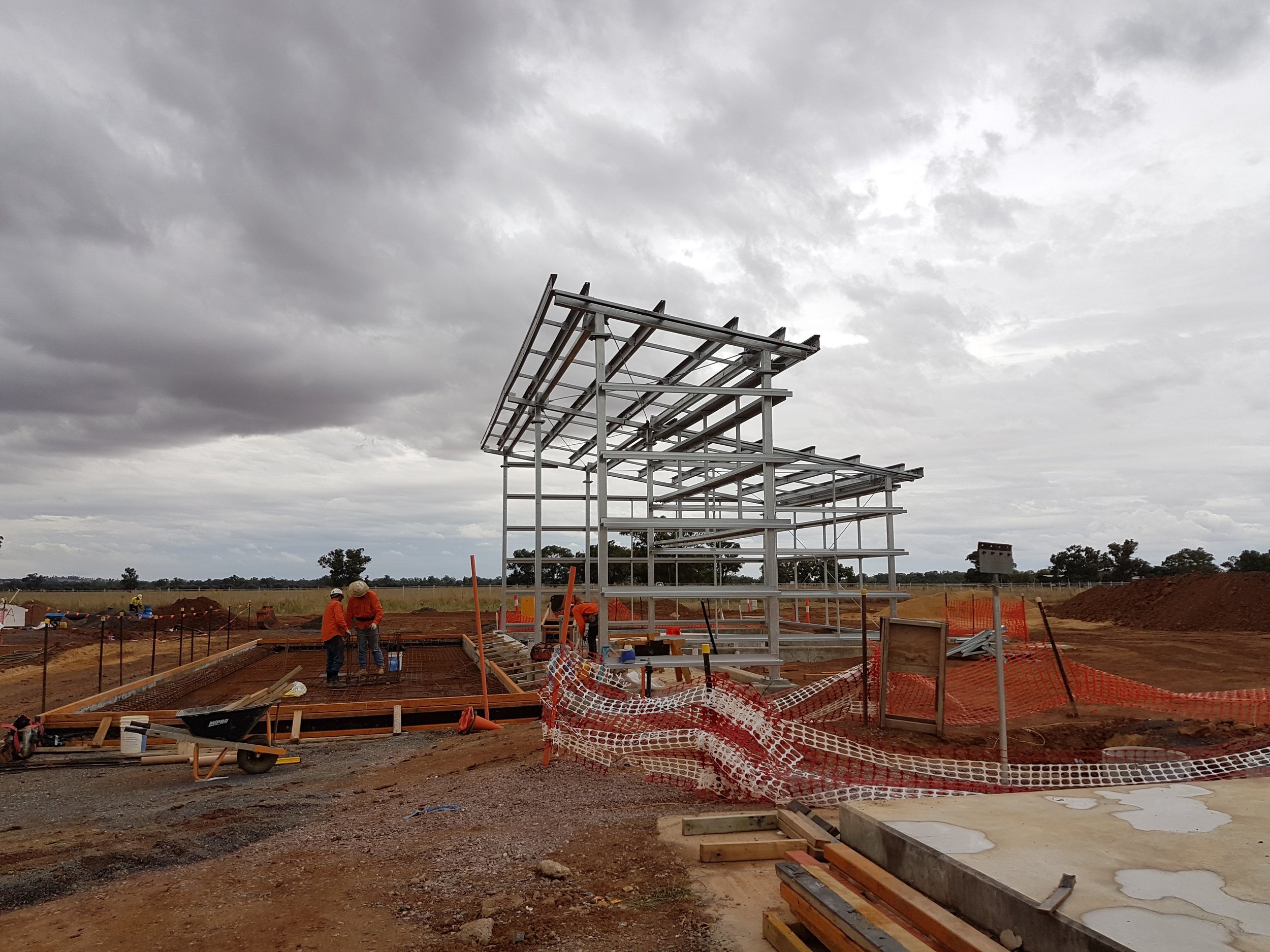 Construction site with a steel framework structure and workers in orange shirts, under cloudy sky.