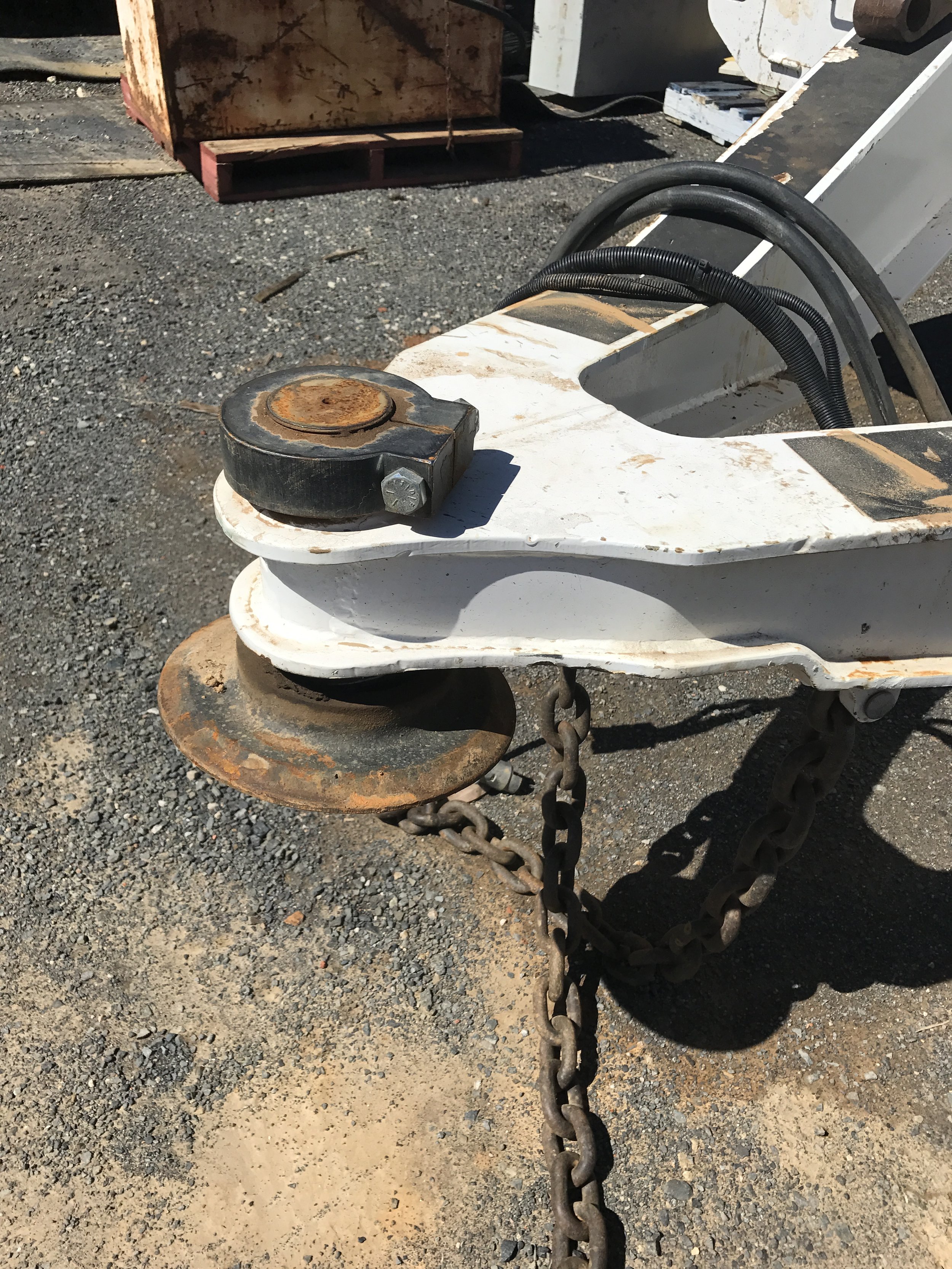Close-up of a rusty metal hook and chain attached to a white metal structure, possibly part of construction equipment or machinery, on a gravel surface.