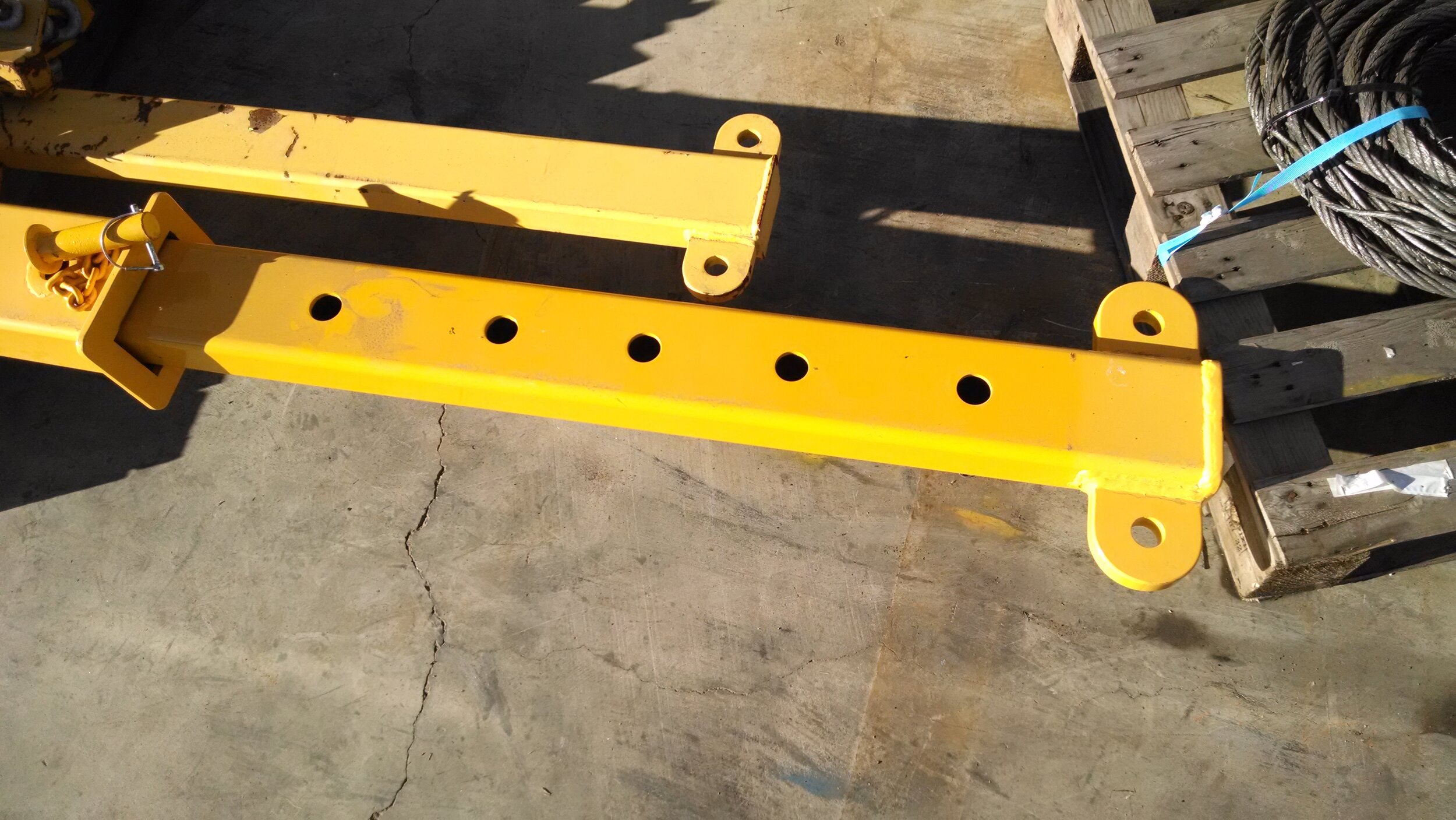 Yellow metal beam with multiple circular holes, part of a construction or industrial equipment, resting on a concrete floor with cracks.