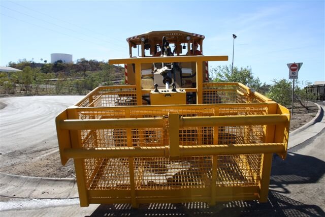 Yellow construction vehicle, likely a road roller, parked on the side of a road with trees and a sign in the background.