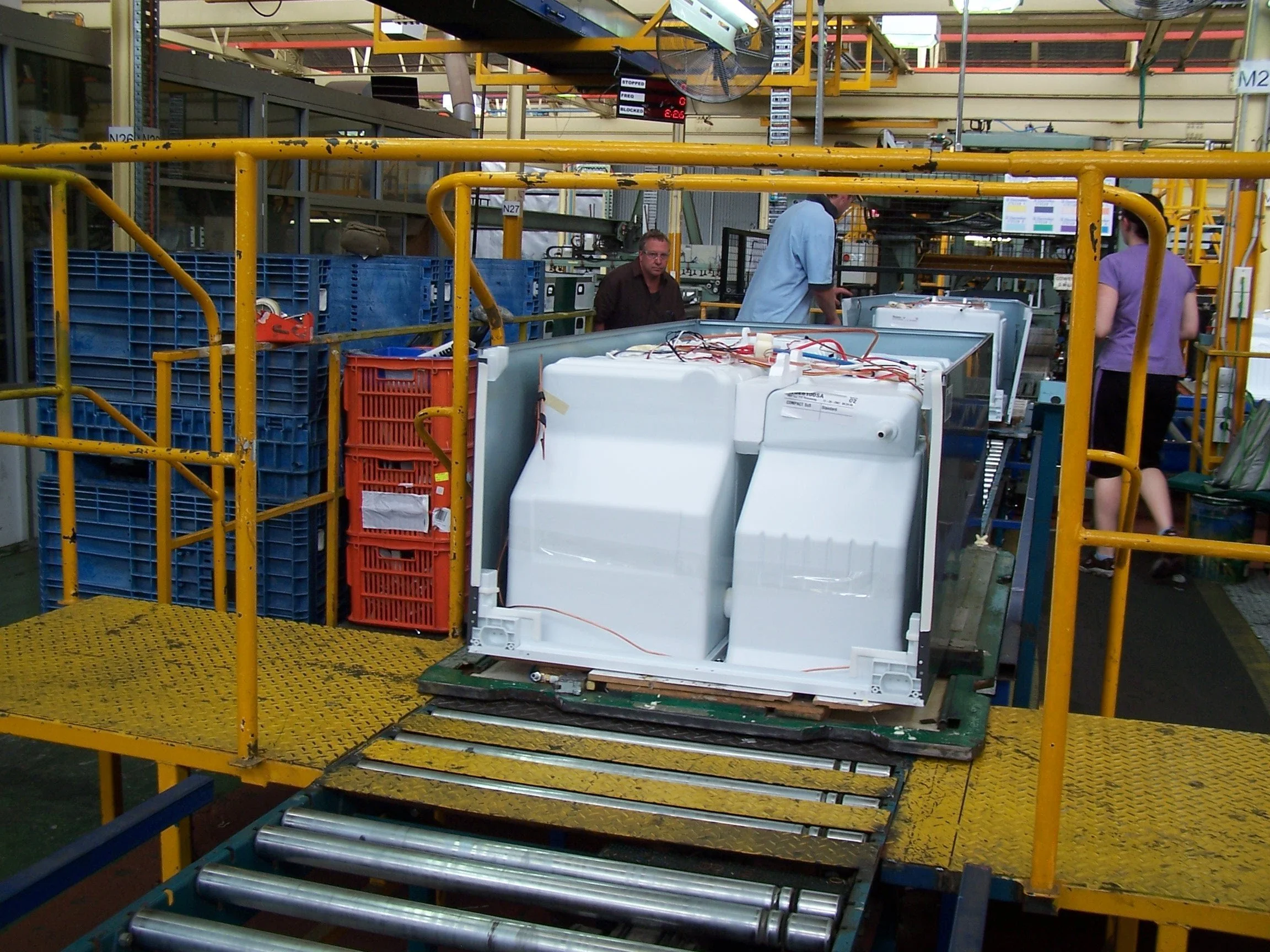 Workers in an electronics manufacturing or repair plant surrounded by yellow safety barriers, machinery, and electronic components.