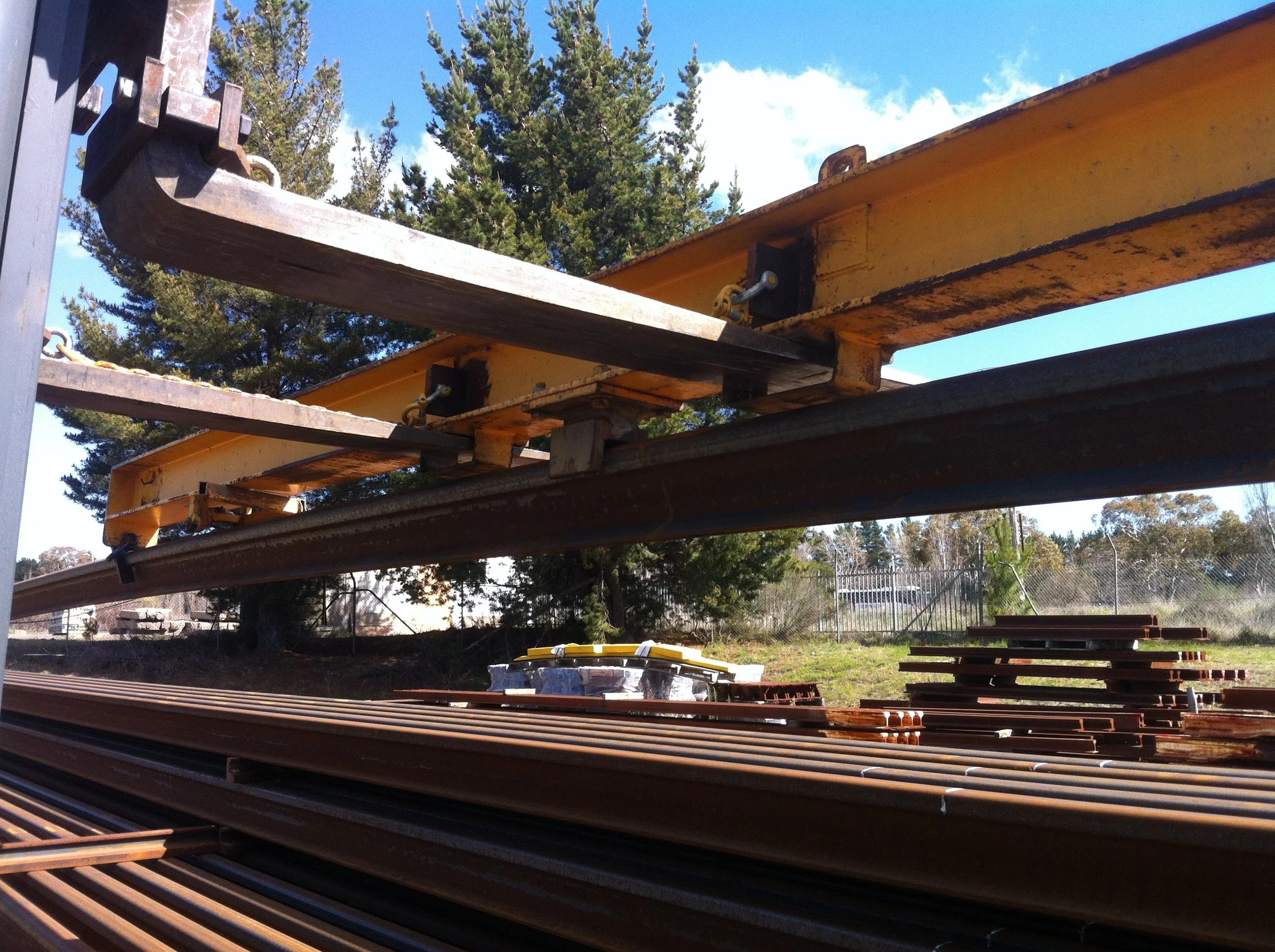 Stacks of construction lumber and metal beams outdoors under a clear blue sky, with trees in the background.
