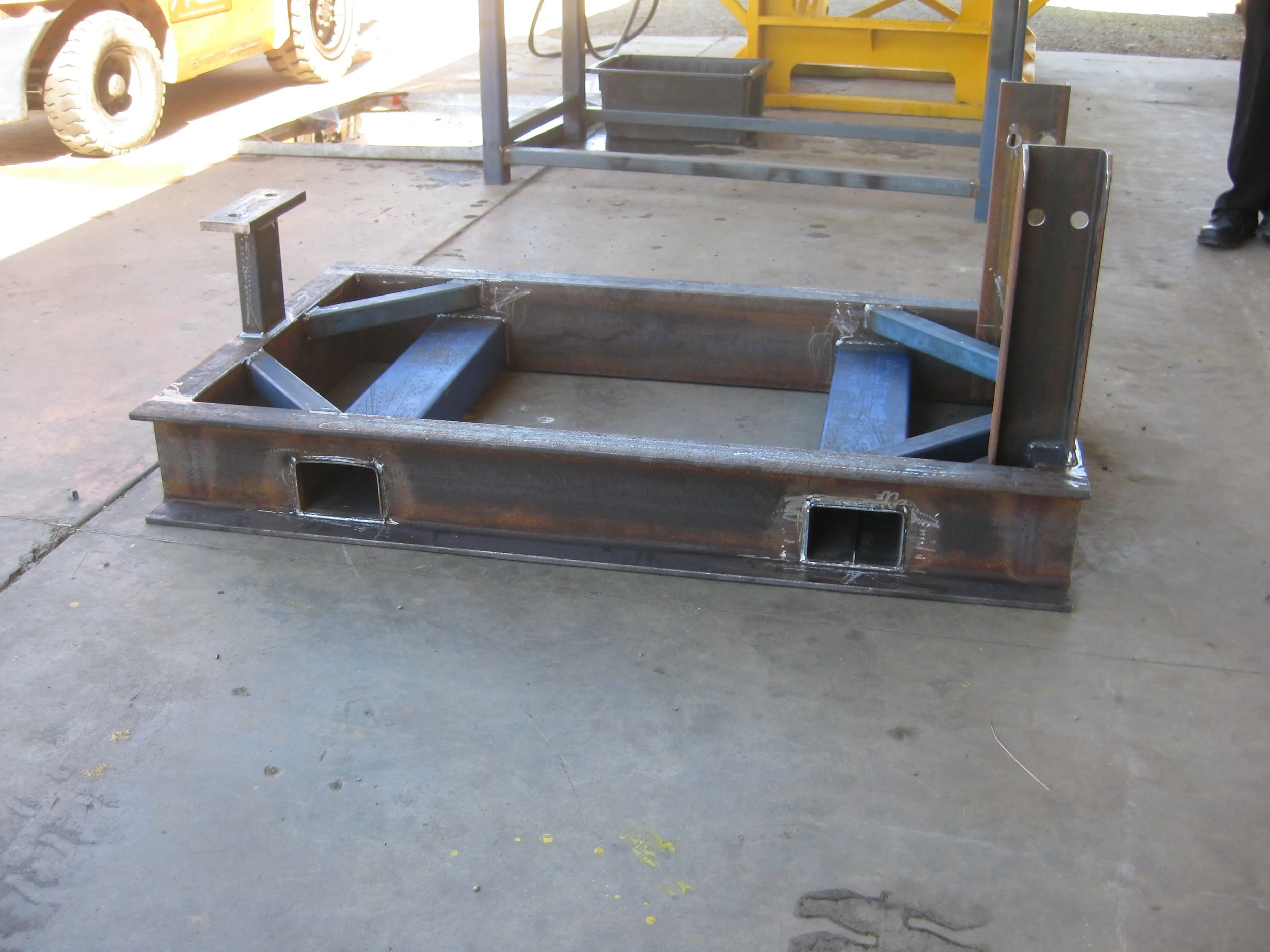 Metal frame structure on the floor of a workshop or garage with a yellow vehicle nearby and a person standing to the right.