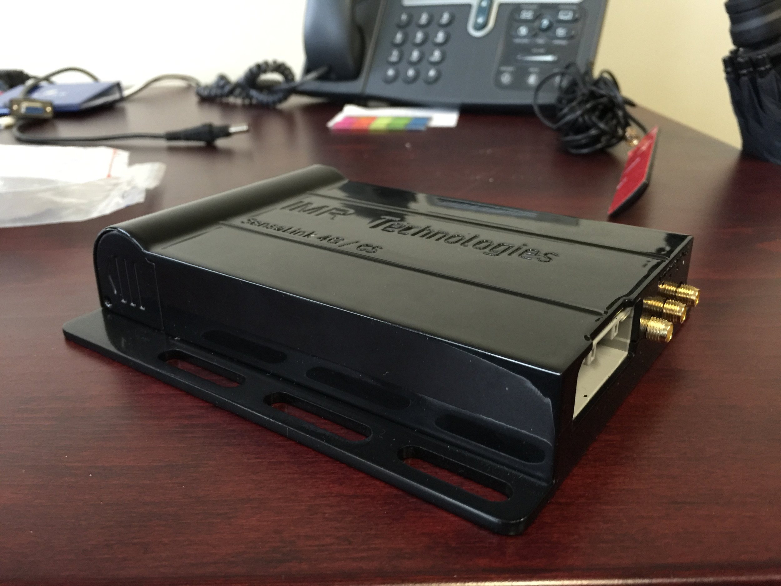 A black TV tuner device with gold connectors on a wooden desk, with a telephone and miscellaneous office items in the background.