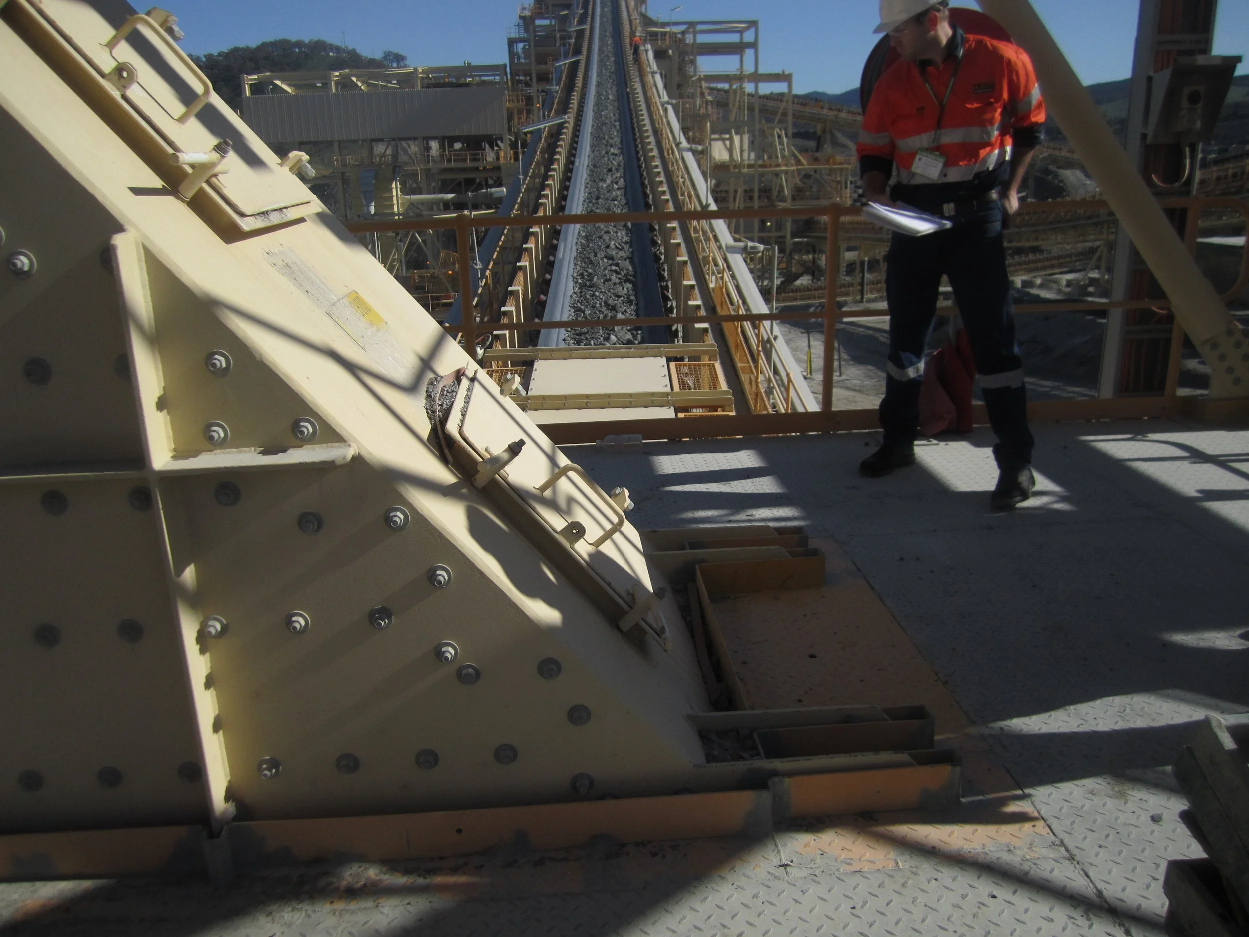 Industrial worker inspecting a large piece of machinery at an industrial facility, with a long conveyor and loading area stretching into the distance.