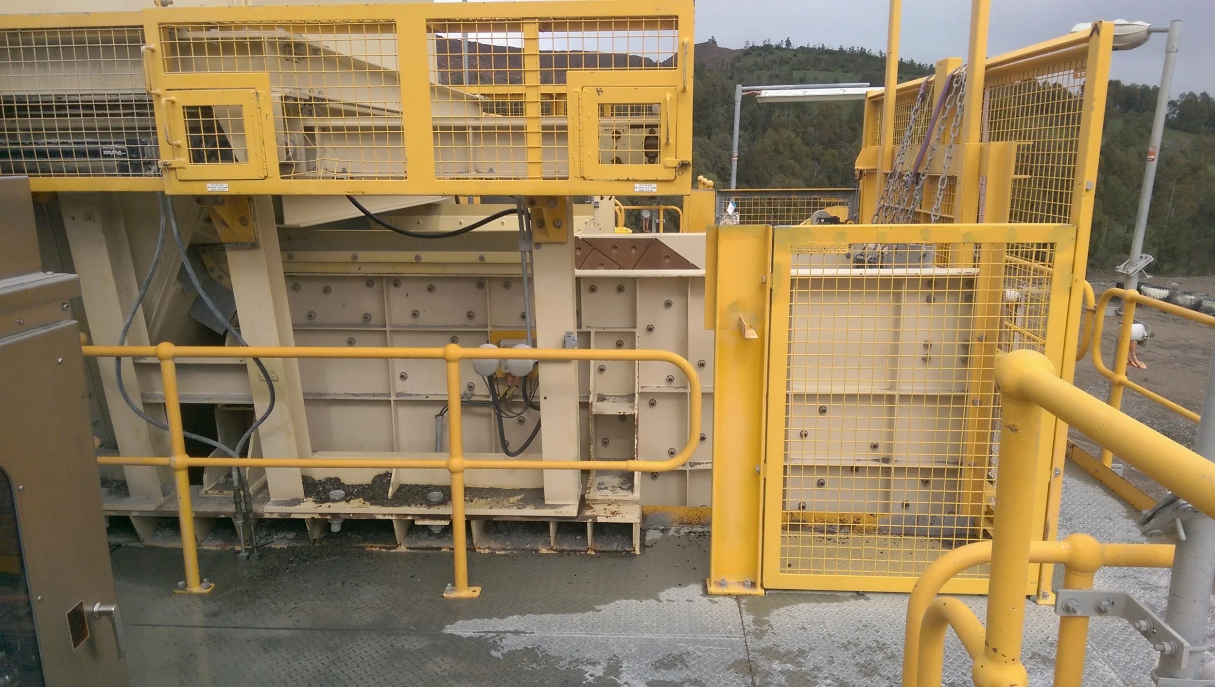 Yellow industrial machinery with safety railings and chains, situated outdoors with hills and trees in the background.