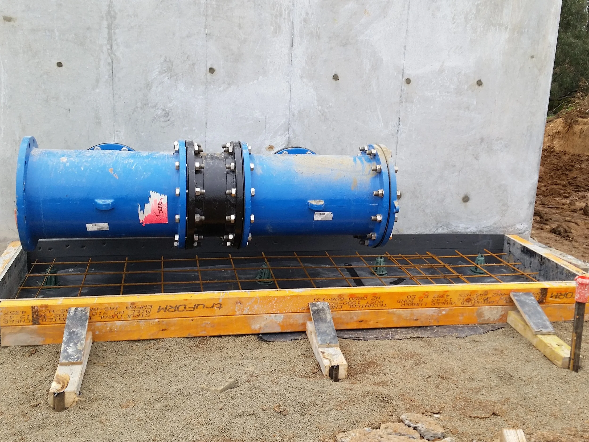 Large blue pipe with flanges and bolts installed on a construction site, mounted on a metal grid within a concrete framework.