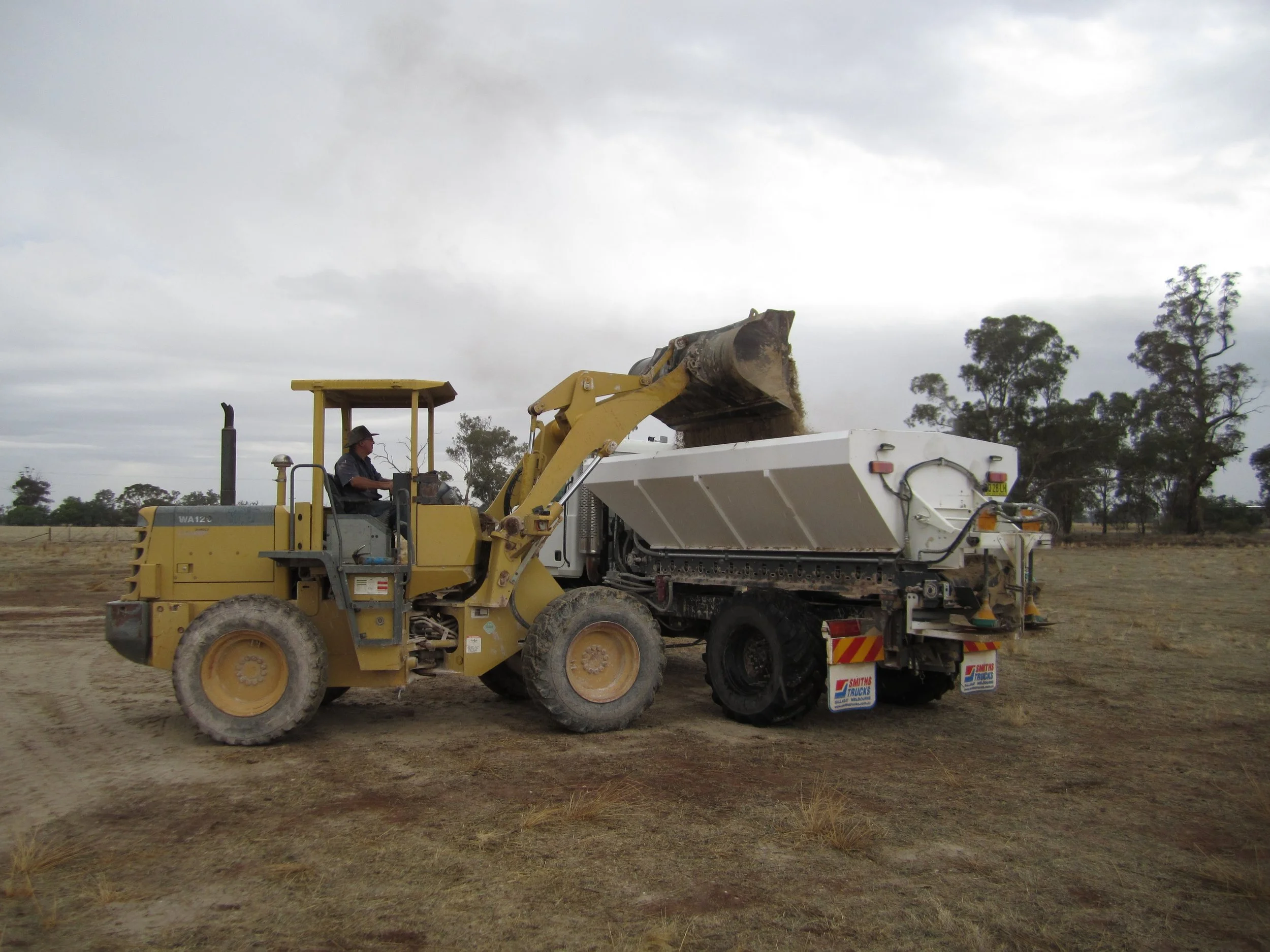A bulldozer loading dirt into a truck on a dry, open landscape with trees and cloudy sky in the background.