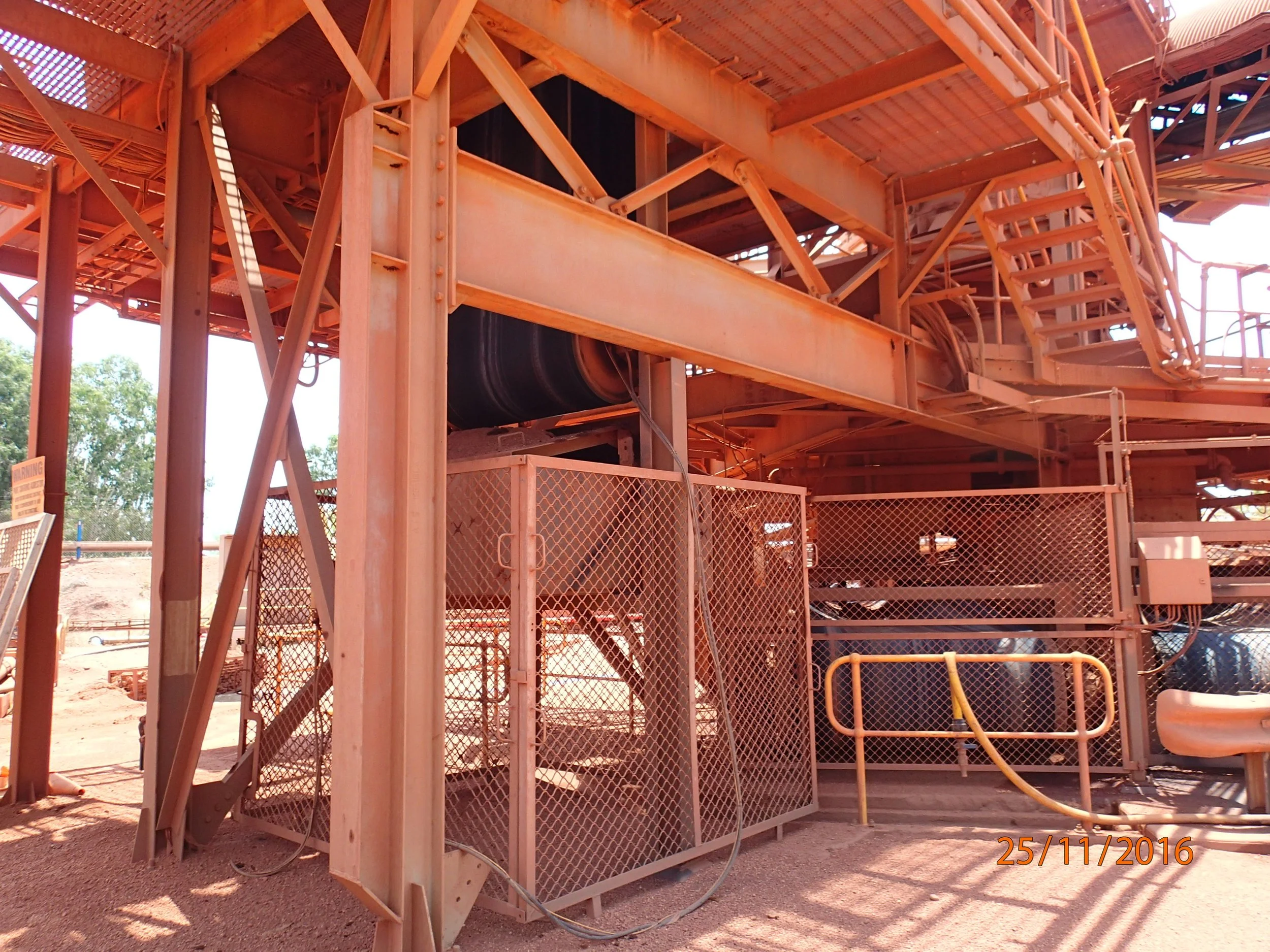 Construction site with wooden scaffolding, stairs, and pipes under a rust-colored metal roof, with trees visible in the background.
