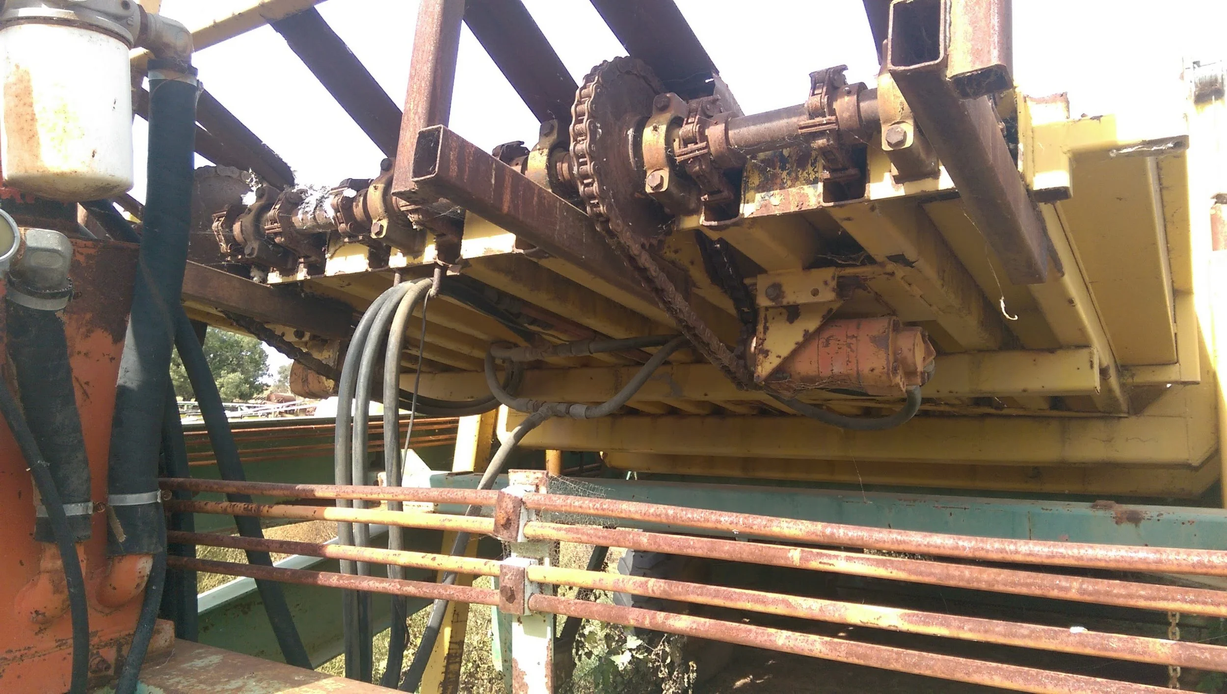 Close-up view of rusted machinery and chains on a yellow industrial machine, with hoses and pipes attached.