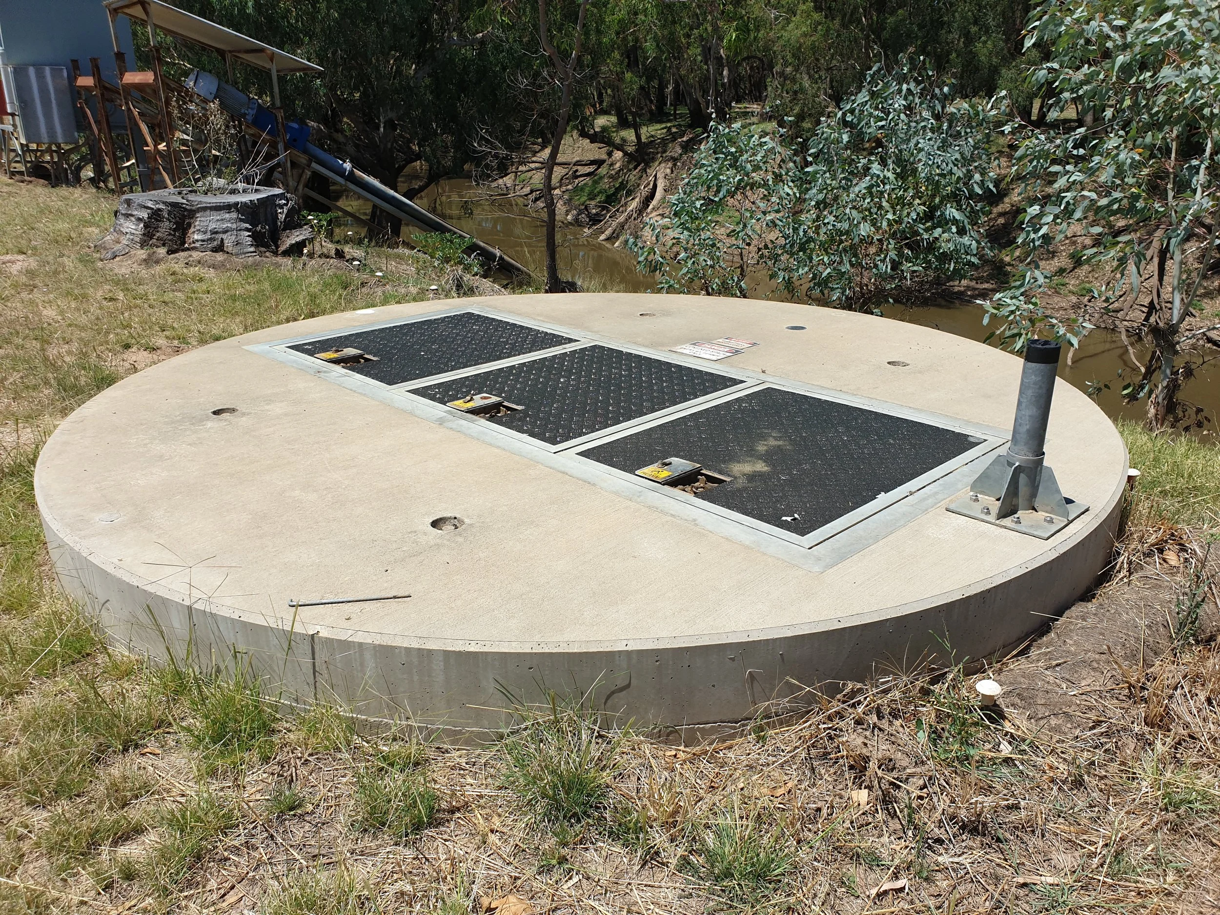A round concrete water tank with access panels and a pipe on a grassy area near a creek with trees.