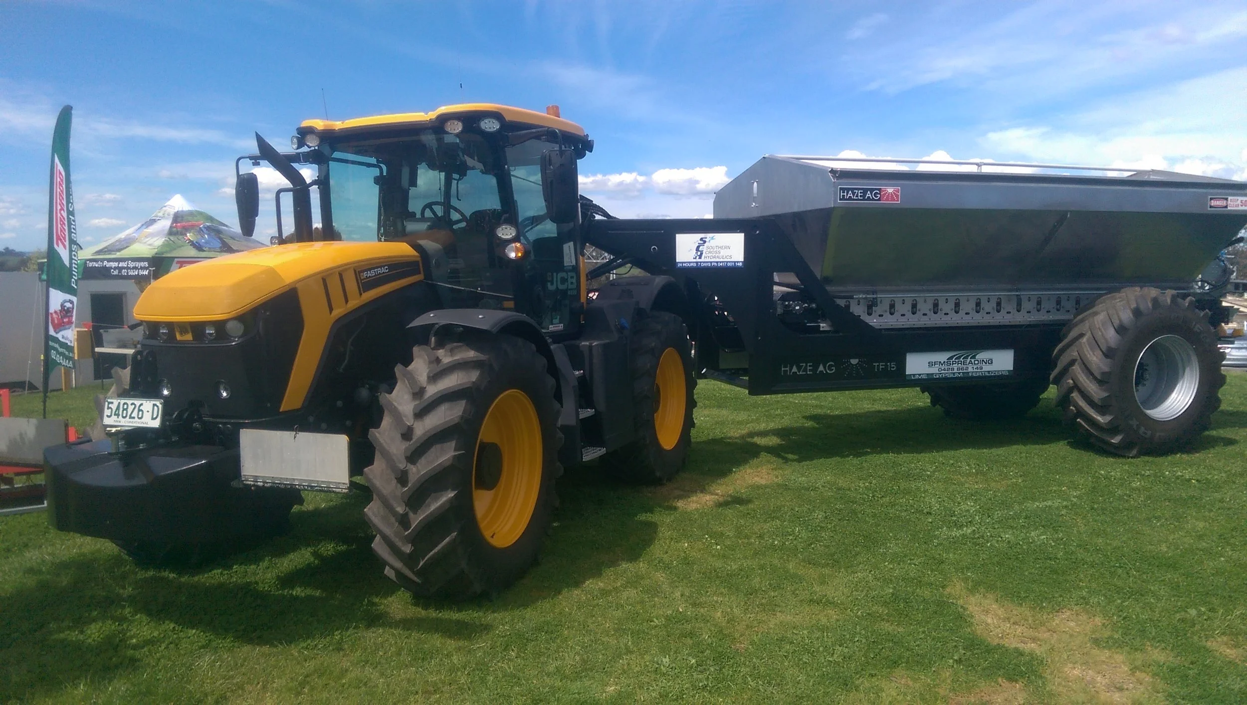A yellow and black tractor pulling a large metal trailer on green grass under a blue sky with clouds.