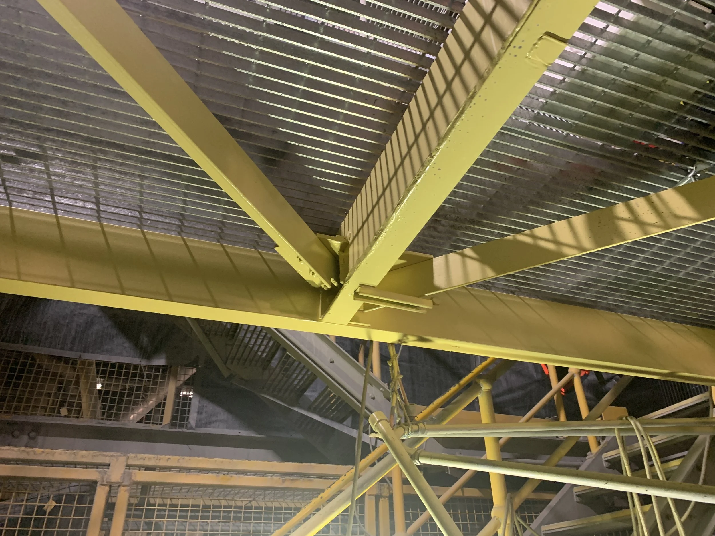 A view looking up at a yellow industrial-style staircase with metal grating steps and supporting beams, set in a dark environment with shadows cast by the structure.