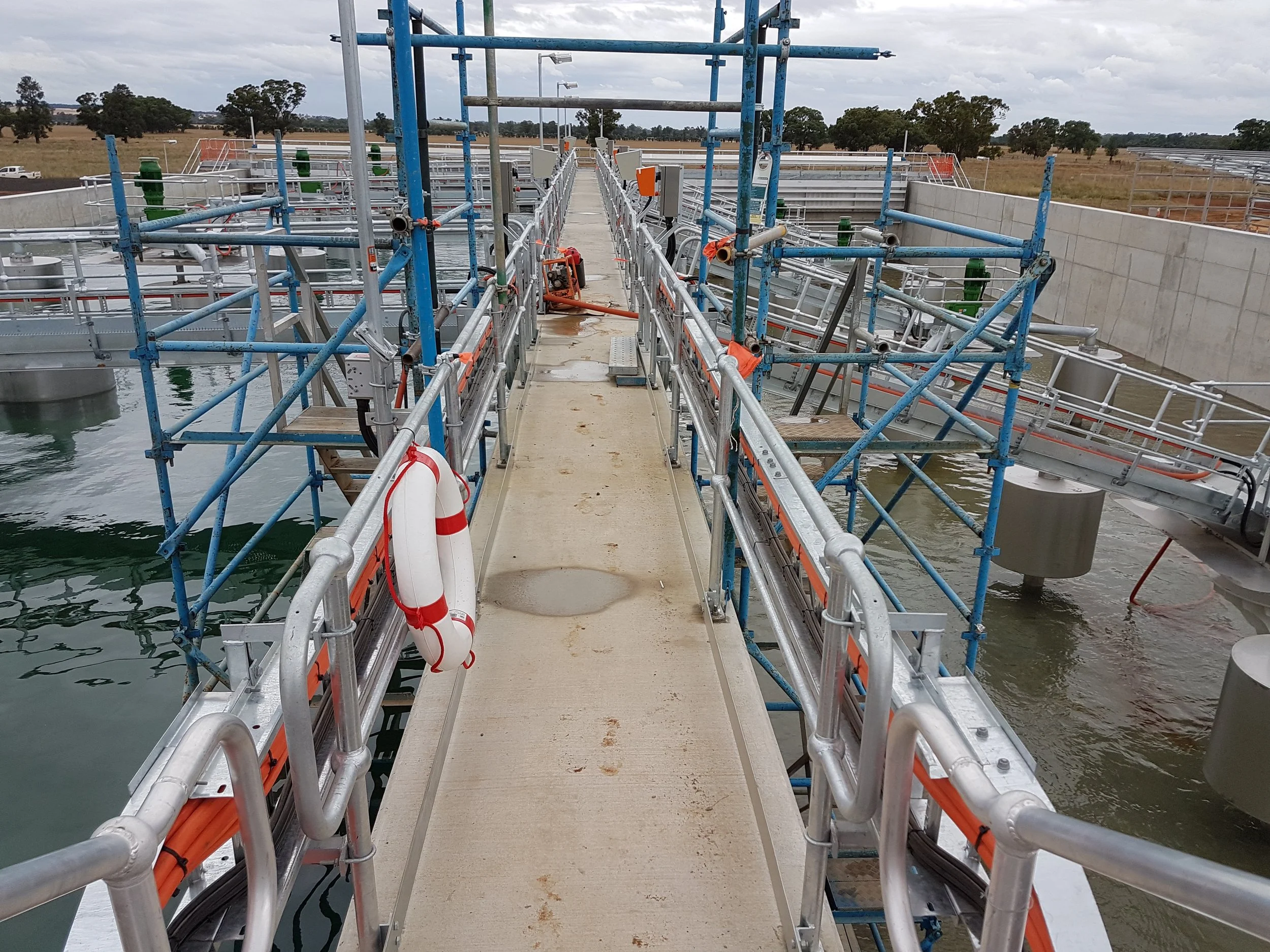 View of a fish farming or aquaculture facility with metal walkways, railings, and scaffolding over water, with pipes and equipment for water and fish management.
