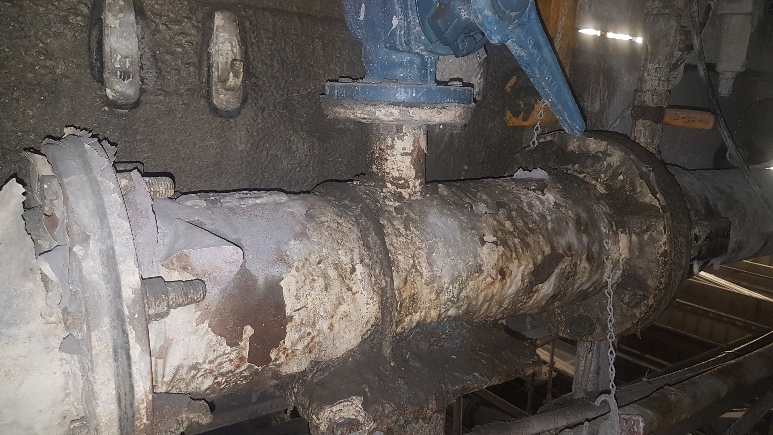 Large, rusted metal pipe or boiler with bolts and chains attached, some broken or chipped parts, in a construction or repair site.